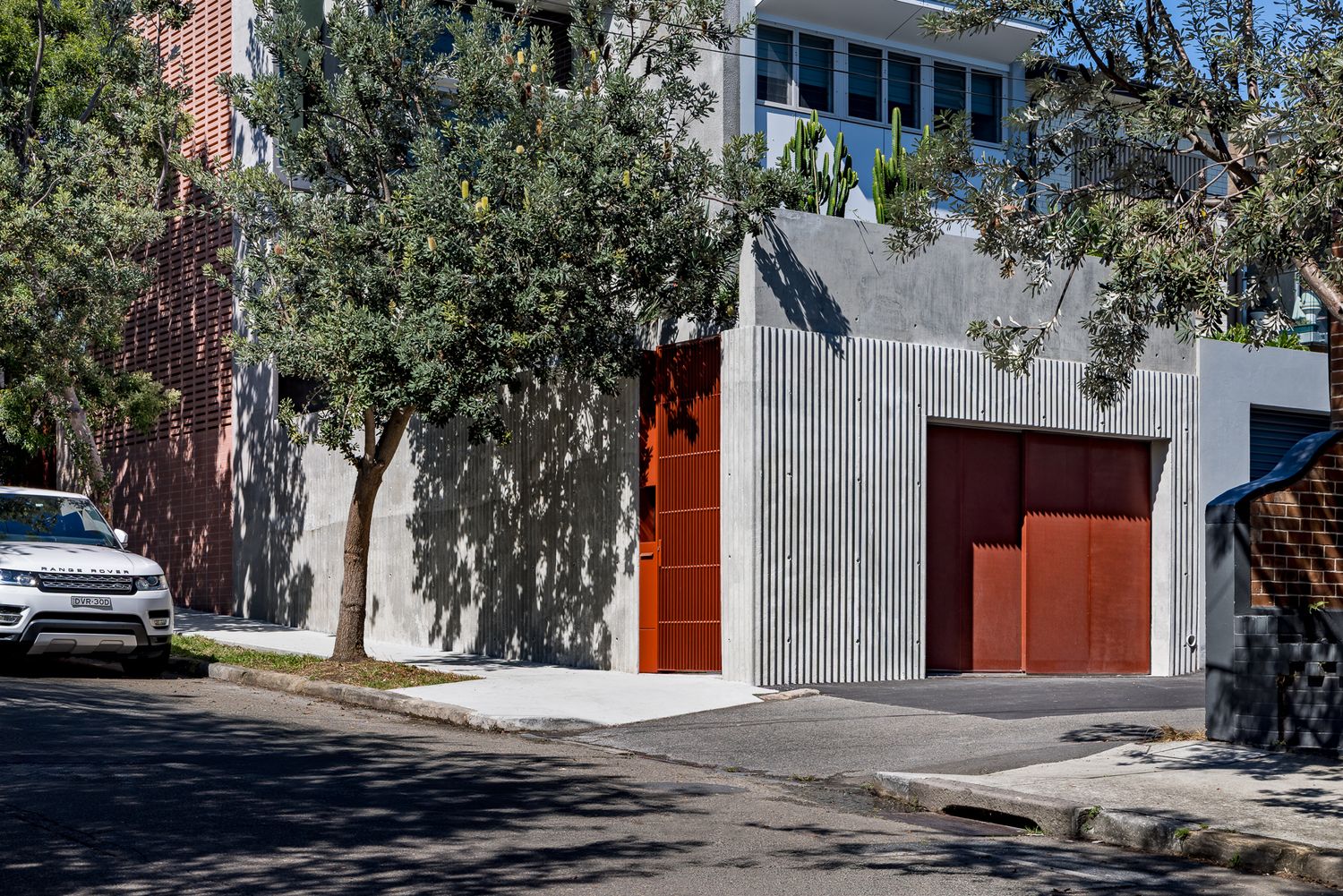 Contemporary concrete house facade on a tree-lined street, featuring timber garage and entry doors and a parked white car.
