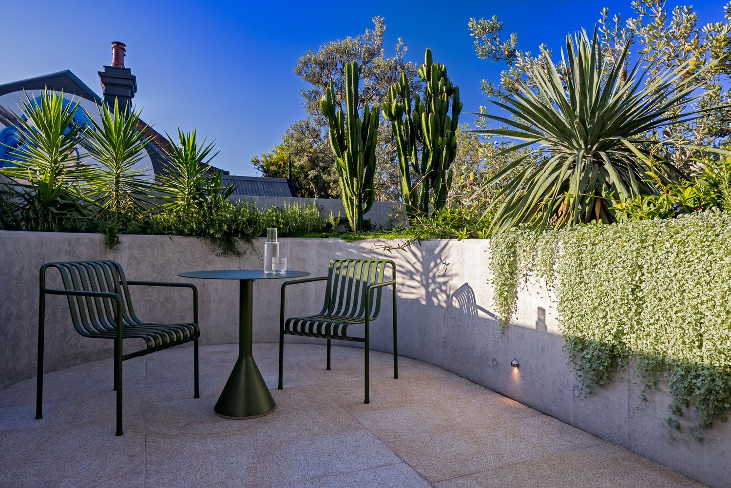 Sunny courtyard patio with a small bistro table, two green chairs, cacti, agave plants, and a curved concrete wall.