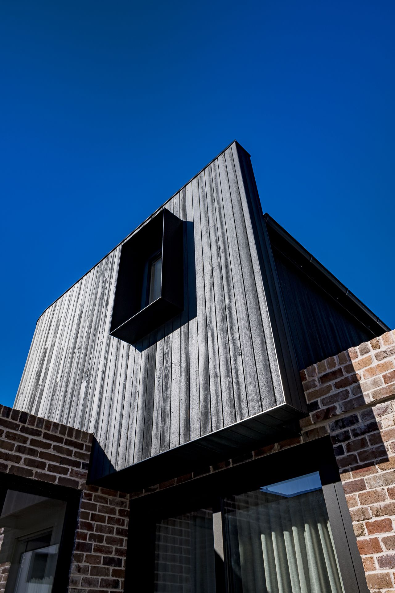 Low-angle view of angular black timber cladding with a recessed window box above rustic brick walls against a deep blue sky.