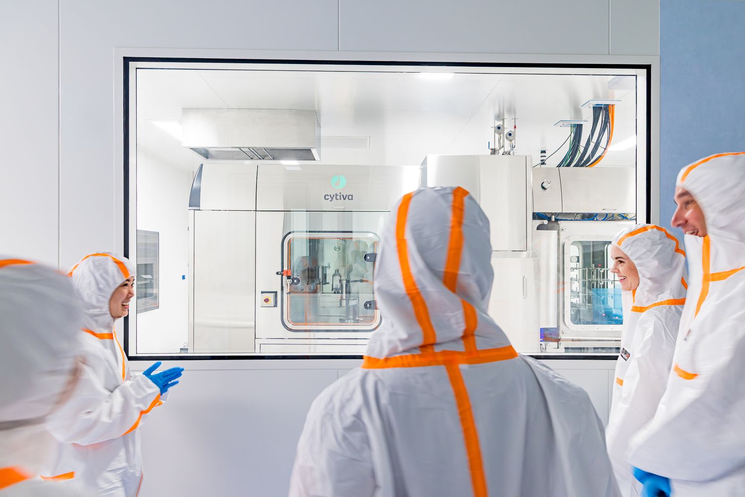 Scientists in white and orange protective coveralls observe bioprocessing equipment through a viewing window in a cleanroom.