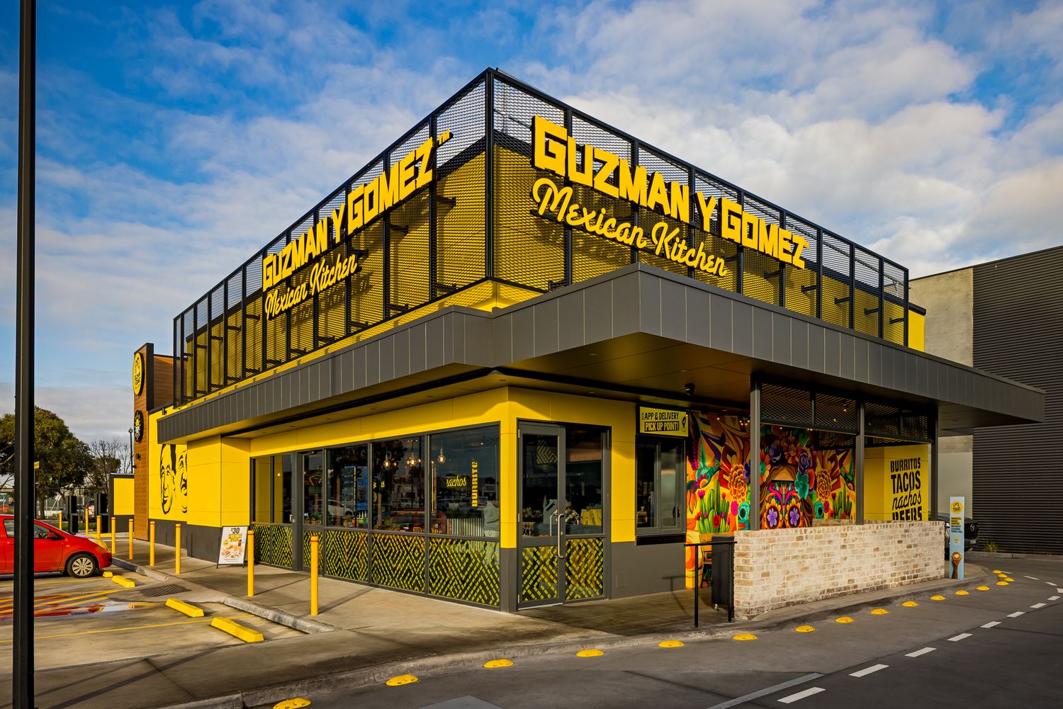 Corner view of a Guzman y Gomez Mexican Kitchen restaurant with yellow signage, large windows and a colorful mural entrance.