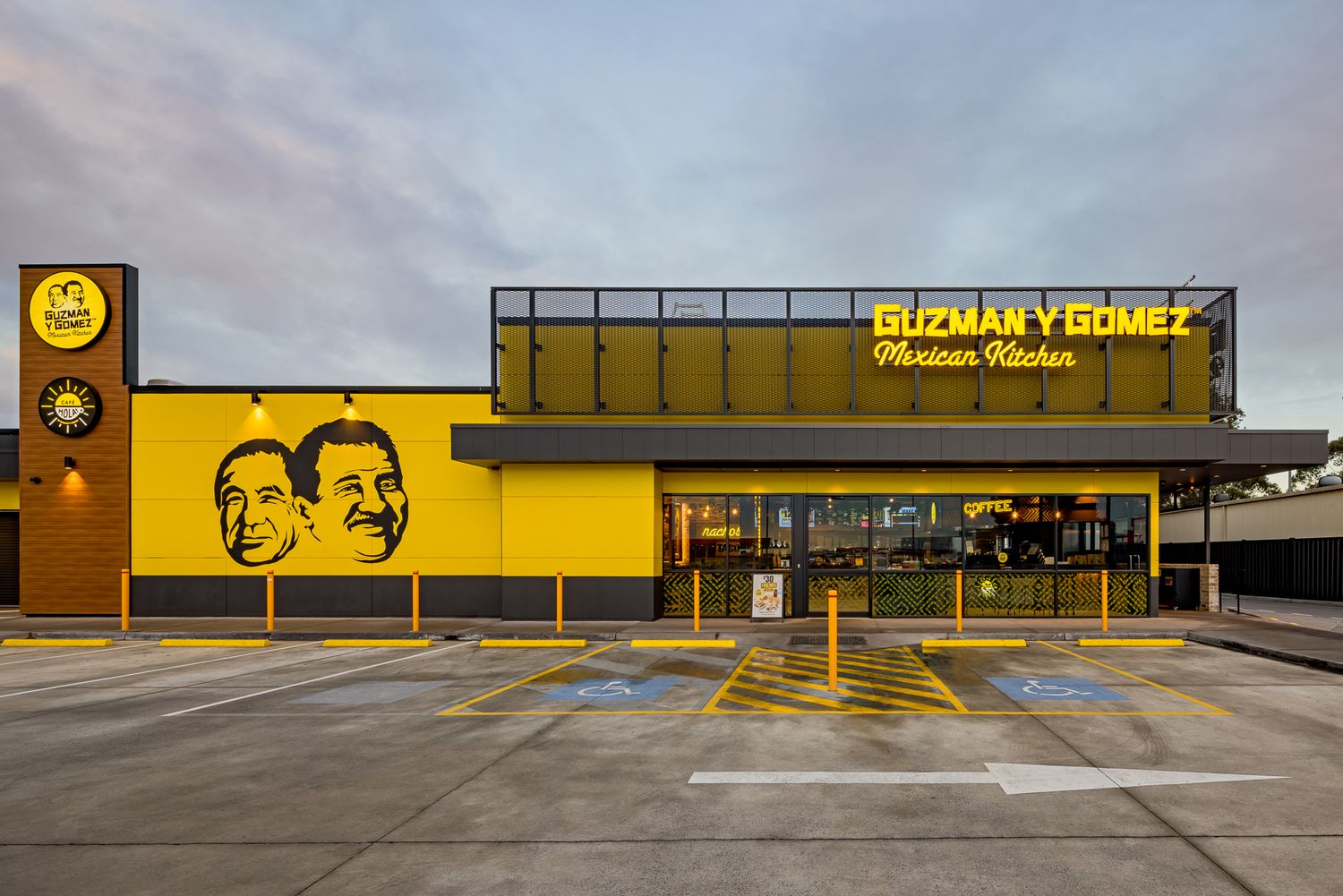Exterior of a yellow Guzman y Gomez Mexican Kitchen restaurant at dusk with a glowing sign and two smiling faces on the facade.