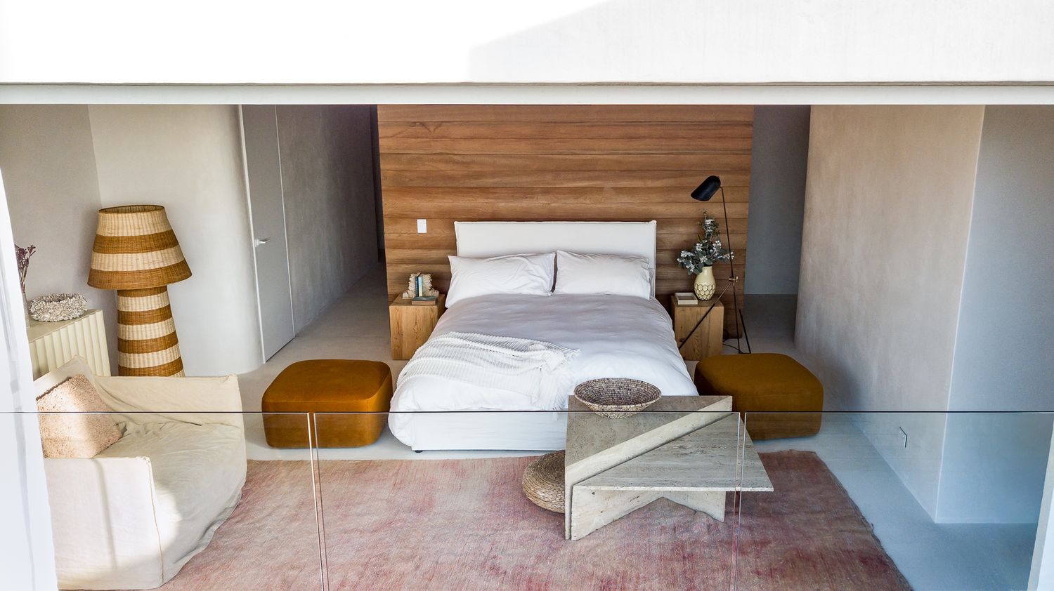 Modern bedroom with a wood-paneled headboard wall, white bedding, mustard ottomans and a sculptural stone coffee table on a pink rug.