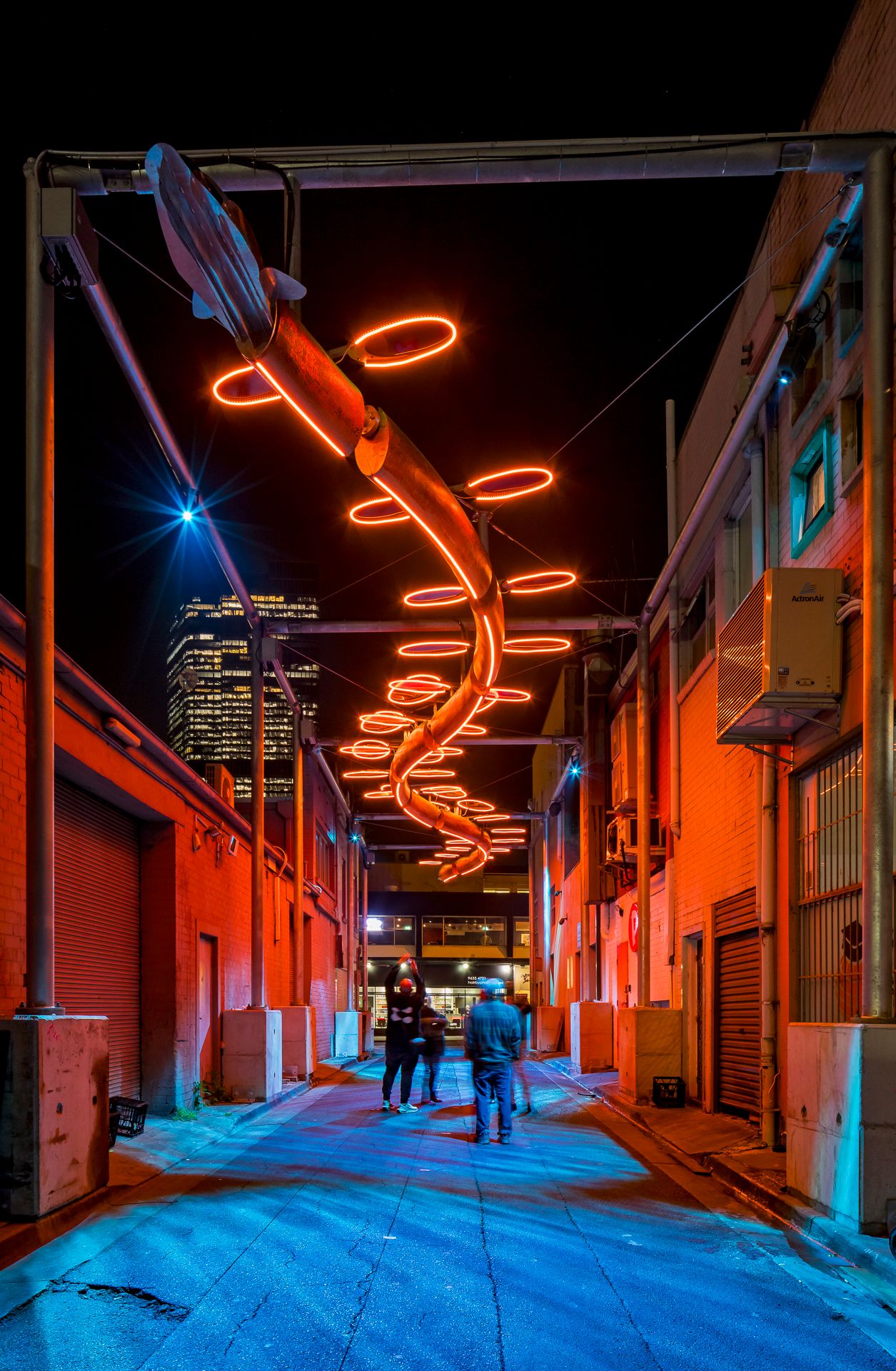 Visitors explore a night-time alley illuminated in warm orange and red by a spiral neon light sculpture suspended above the walkway.