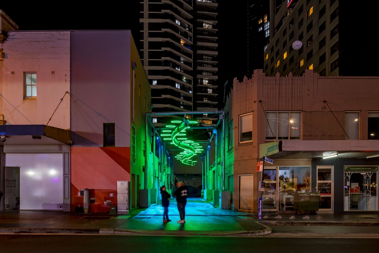 Green neon spiral light sculpture glows over a laneway entrance at night while two people stand on the teal-lit pavement.