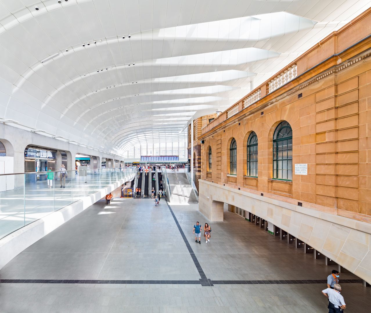 Railway station concourse where a sweeping white ribbed roof meets a heritage sandstone building with arched windows.