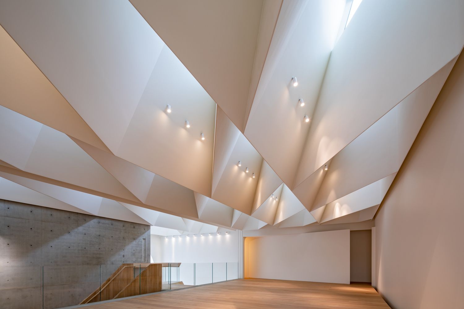 Light-filled gallery space with folded white angular ceiling panels, skylights, timber flooring and a concrete wall.
