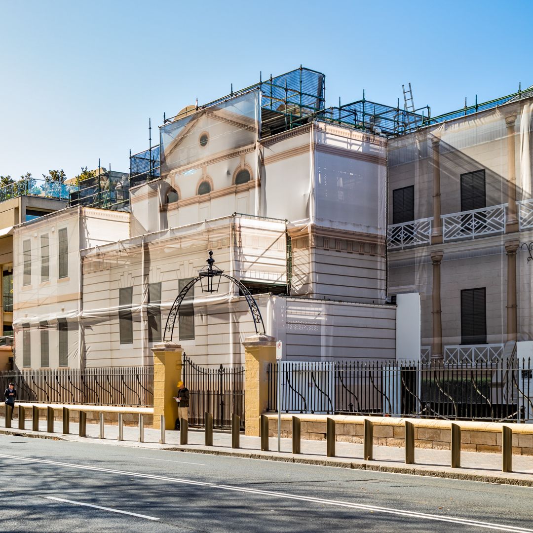Historic cream-colored building wrapped in scaffolding and protective sheeting during renovation behind a black iron fence.