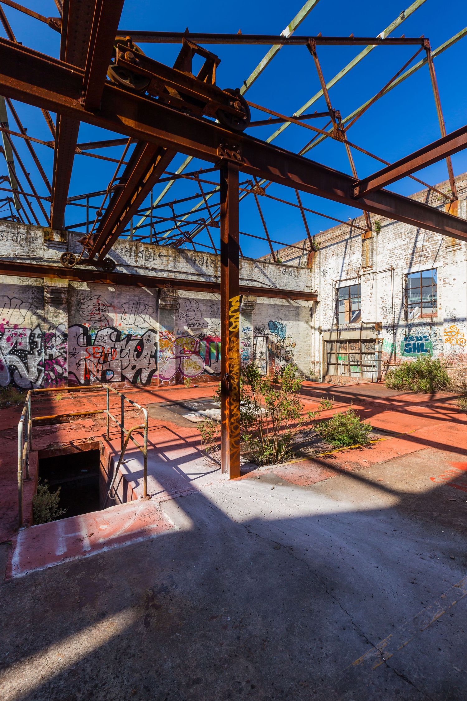 Derelict industrial building with exposed rusted steel beams, graffiti-covered walls and weeds growing under a bright blue sky.