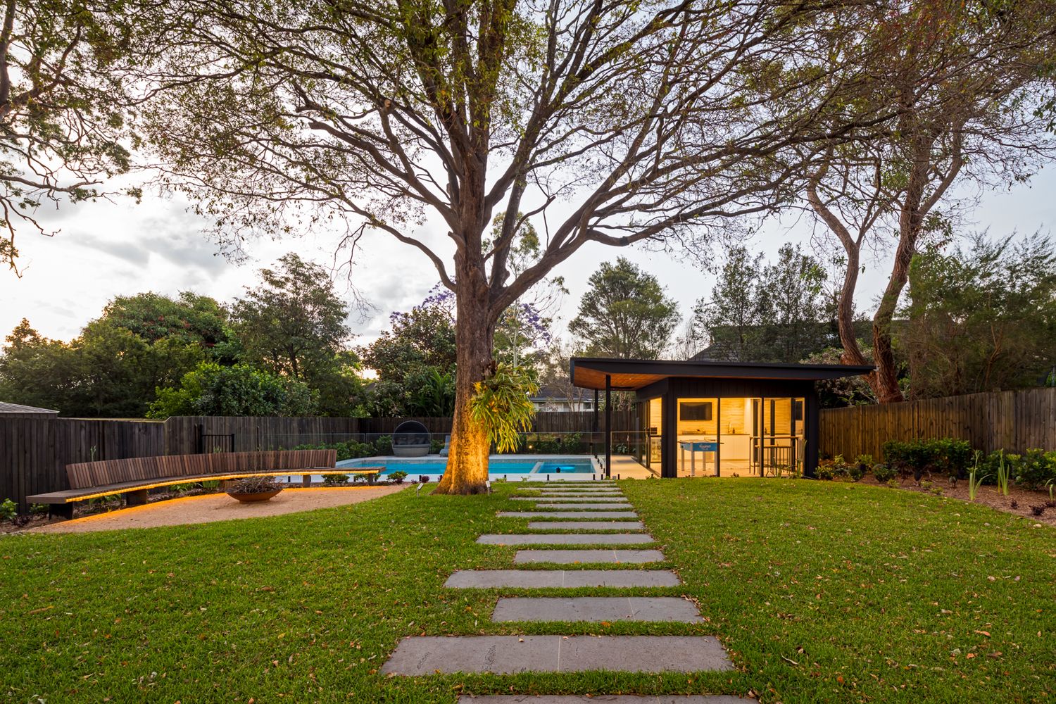 Stepping stone path crosses a green lawn toward a modern garden pavilion and swimming pool beneath large spreading trees.