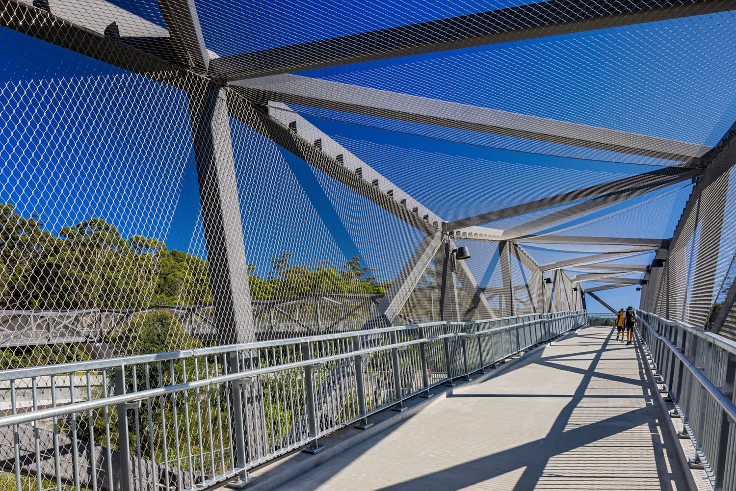 Pedestrian footbridge with angled steel trusses and blue mesh screens running through bright sunlight and shadow above leafy trees.