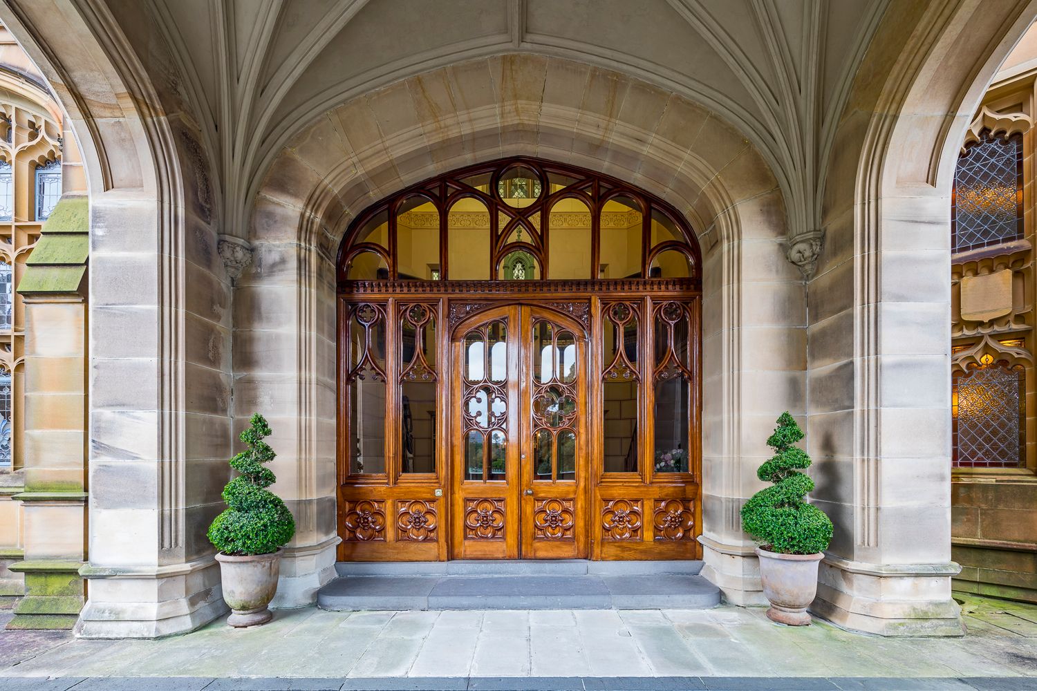 Ornate timber double doors with stained glass set in a vaulted sandstone archway, flanked by potted topiary shrubs.
