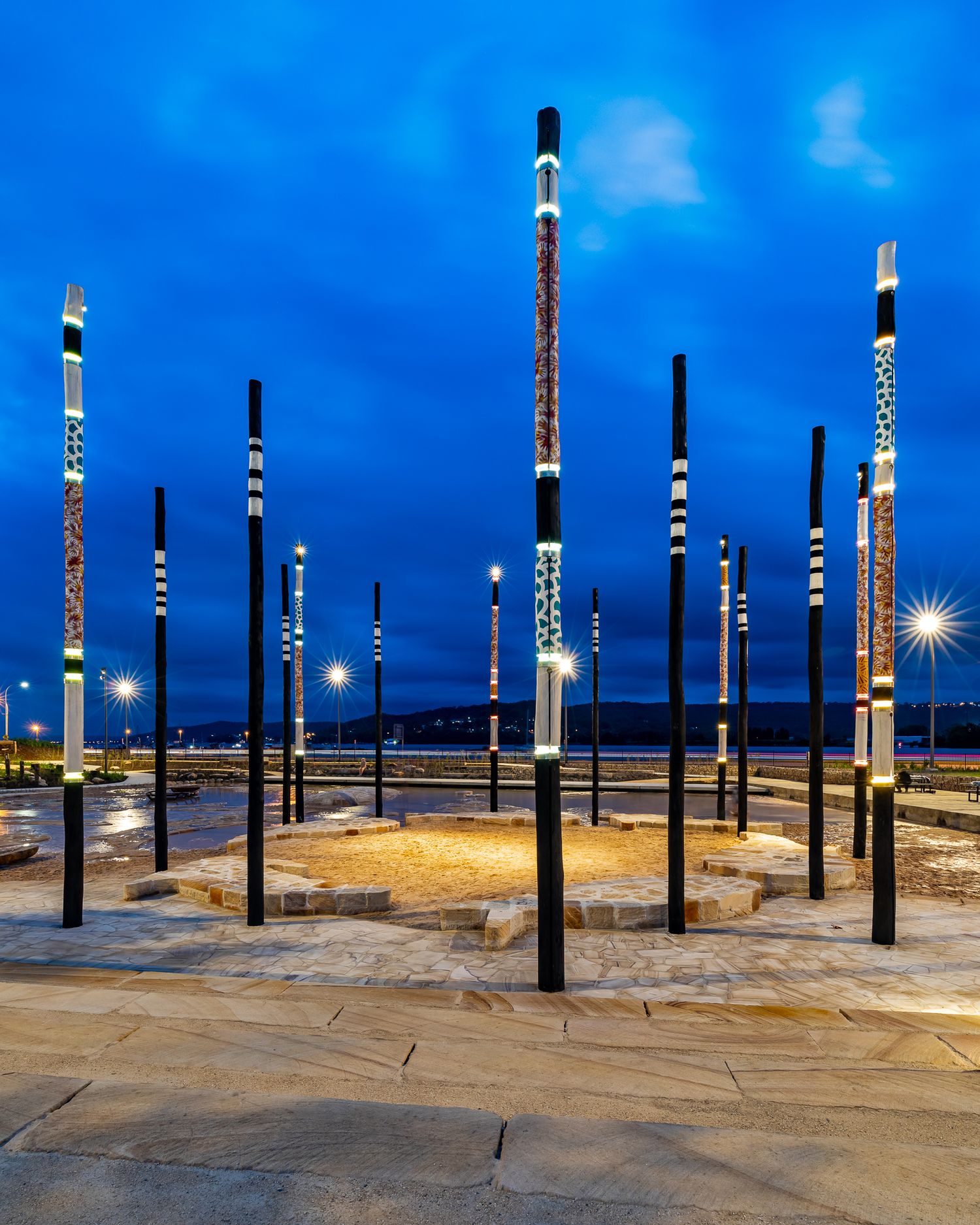 Ring of illuminated art poles with decorative patterns stands around a sandy circular plaza under a vivid blue twilight sky.