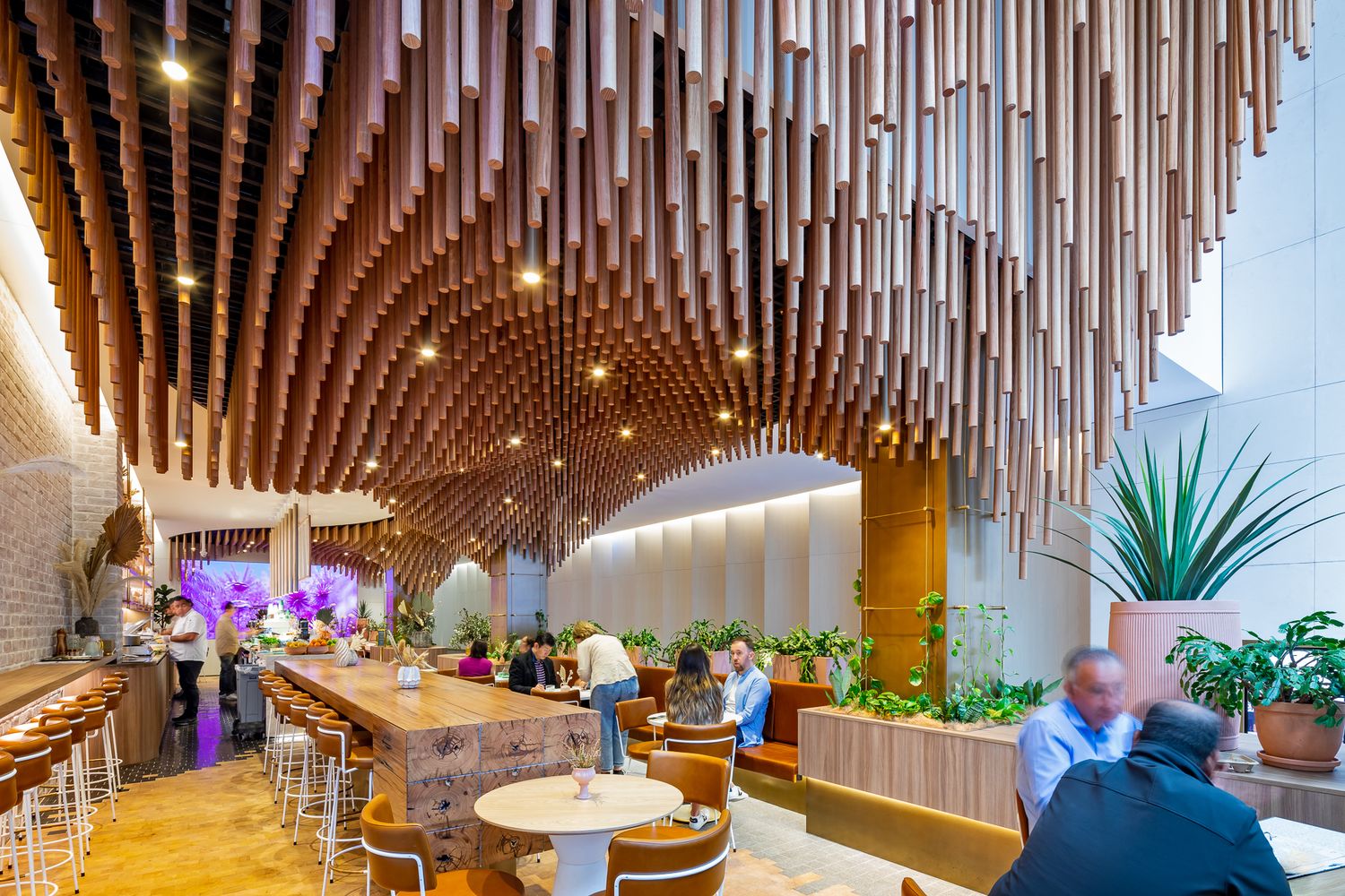 Customers sit at tables and a long communal counter beneath a dramatic cascading wooden baton ceiling in a bright, plant-filled cafe.