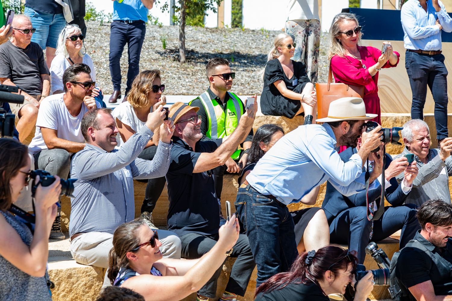 Crowd of seated spectators at an outdoor event taking photos and video with cameras and phones on a sunny day.