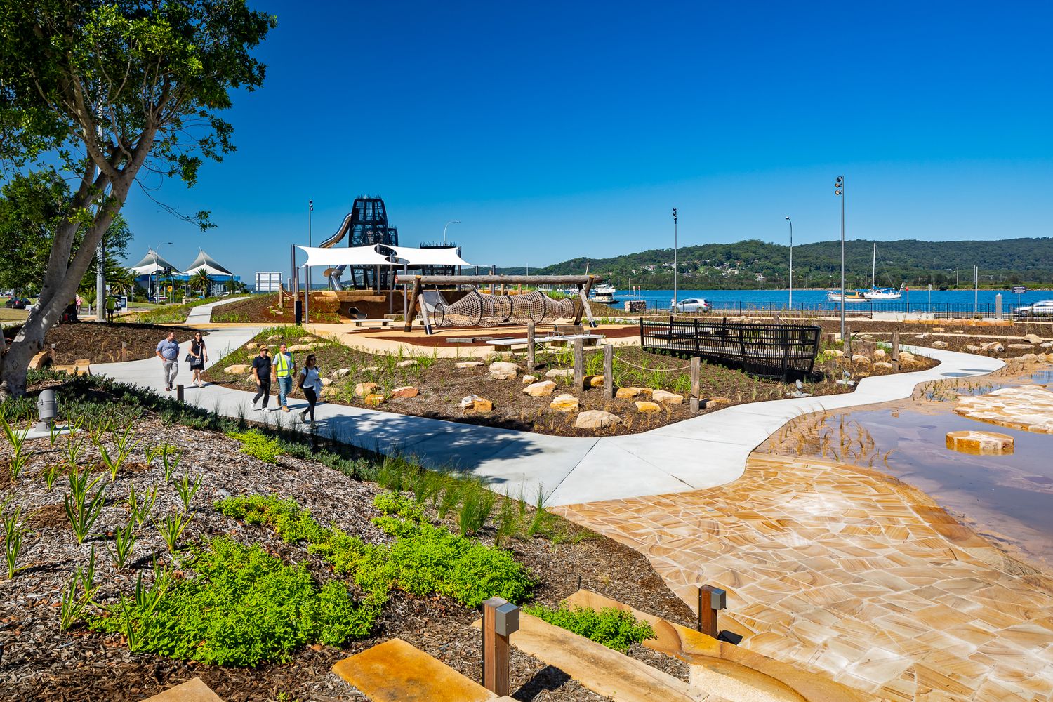 Curving pathway and timber boardwalk through a landscaped waterfront park with a pavilion, visitors and boats on the bay.