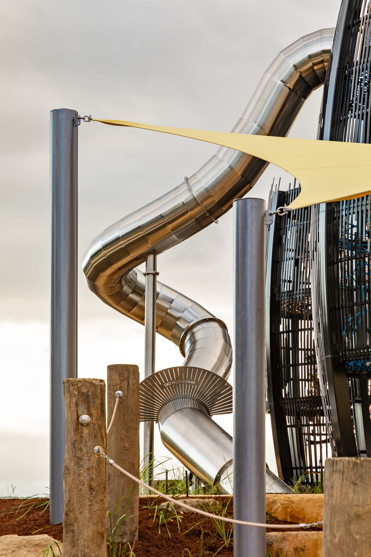 Shiny stainless steel spiral slide twisting beside a black mesh climbing tower, with a yellow shade sail and timber posts.