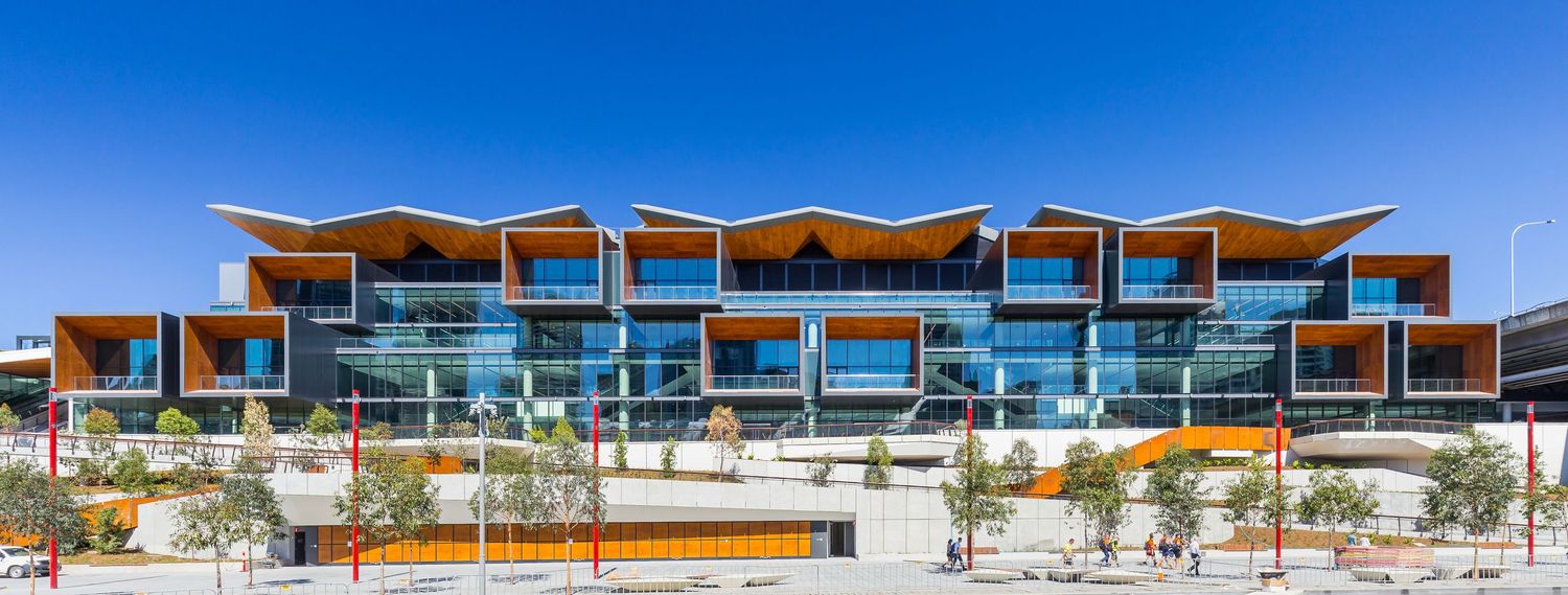 Modern waterfront convention centre facade with stepped timber-lined balconies, glass walls and landscaped planting under a bright blue sky.