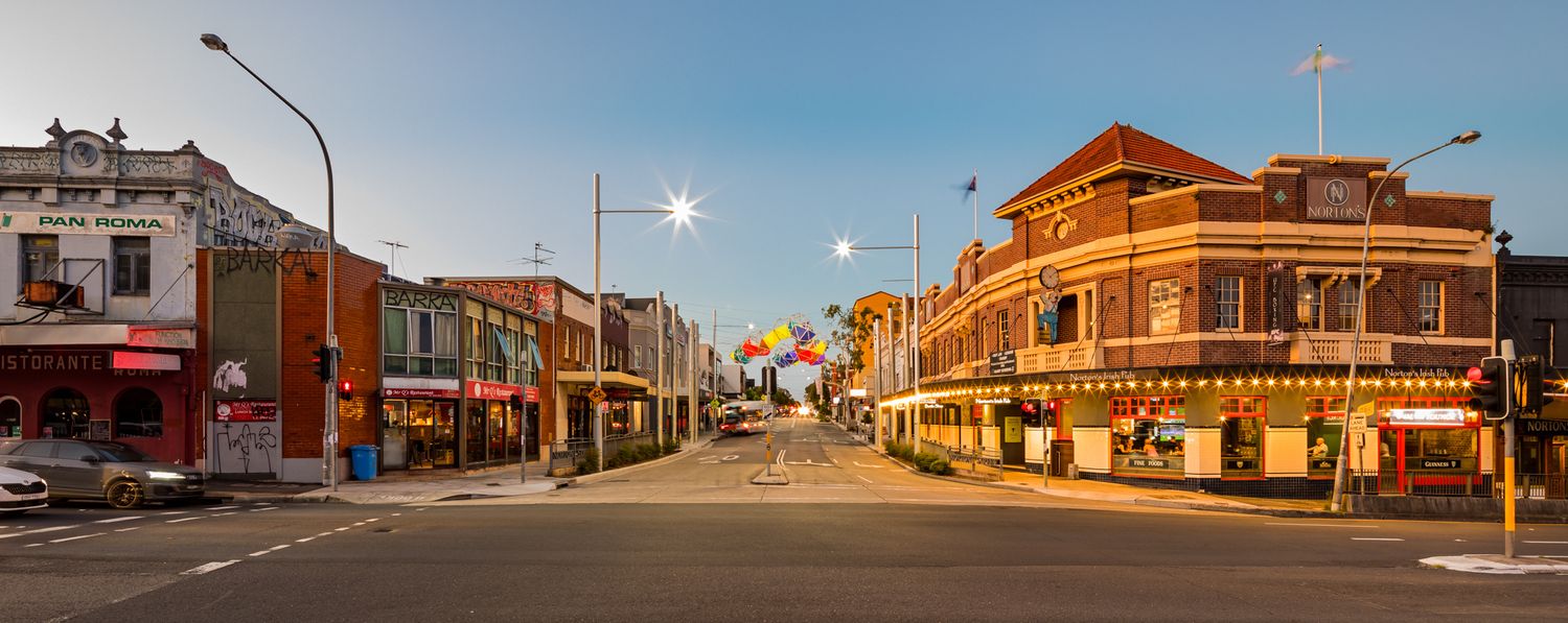 Historic brick corner buildings with shopfronts along a quiet street at dusk with warm streetlights glowing.