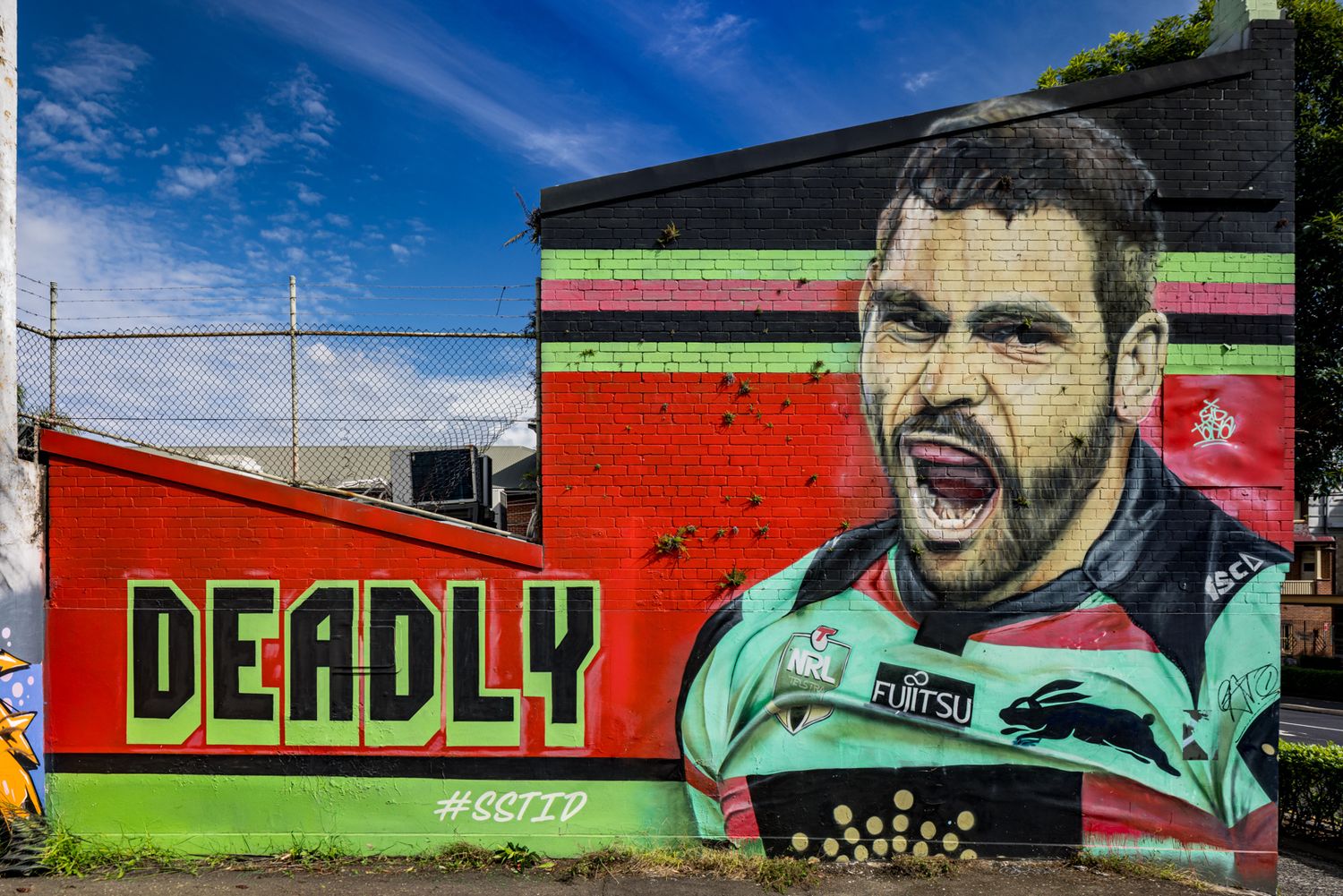 Colorful street art mural of a bearded rugby player with mouth open beside the word DEADLY on a red brick wall.