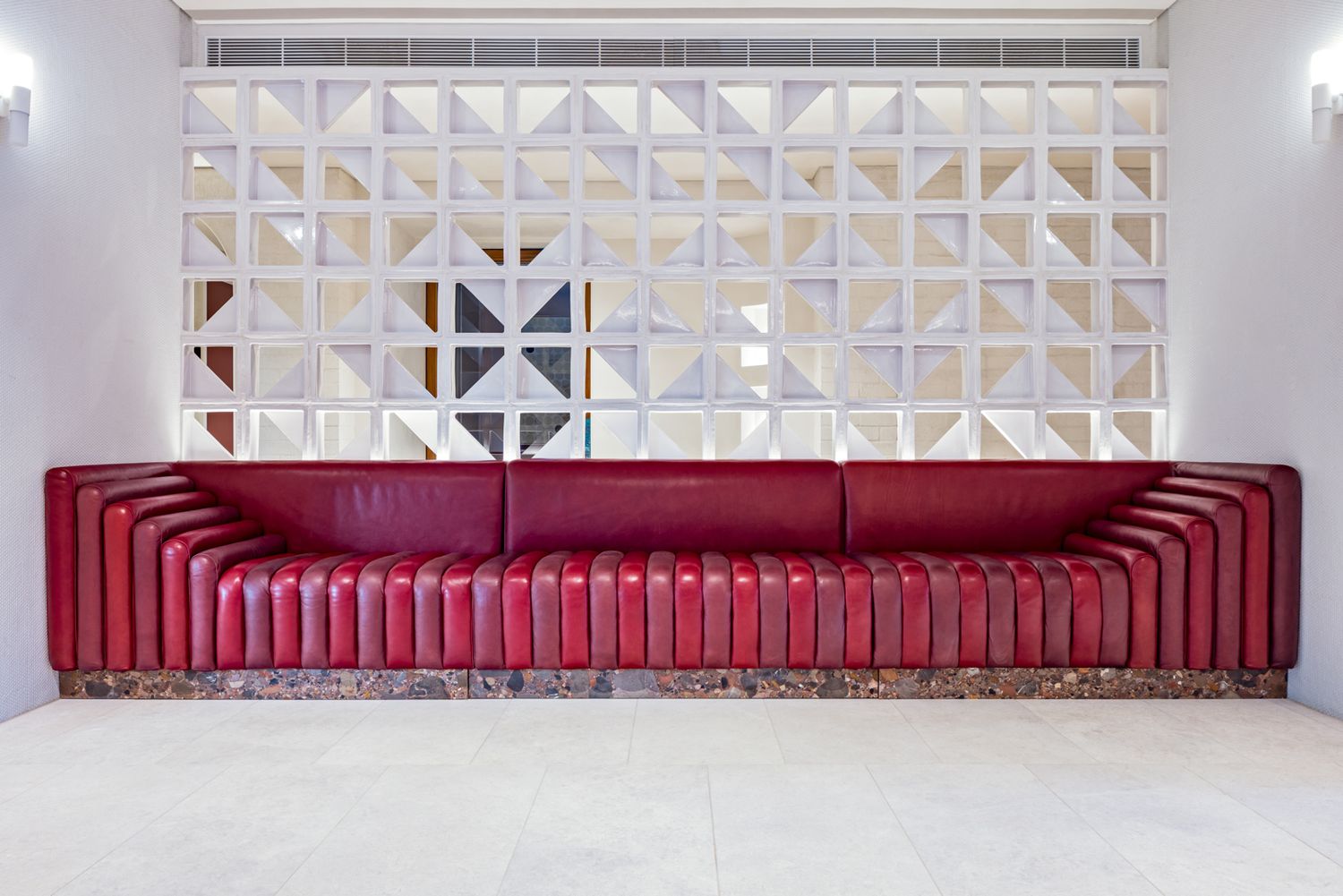 Long red tufted leather banquette in front of a white geometric breeze block screen wall in a bright, modern interior space.