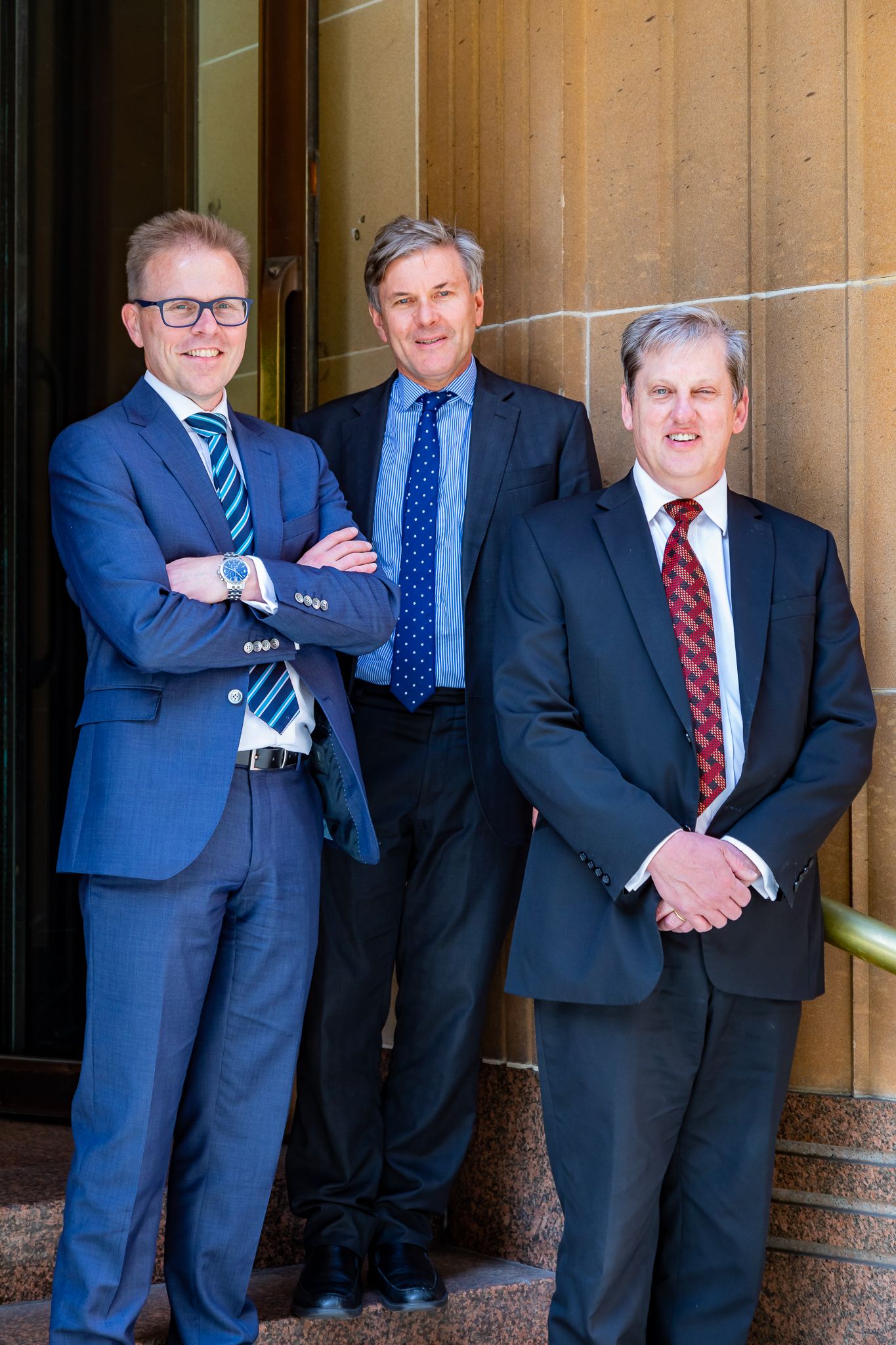 Three men in business suits and ties standing together on stone steps in front of a sandstone building, smiling at the camera.