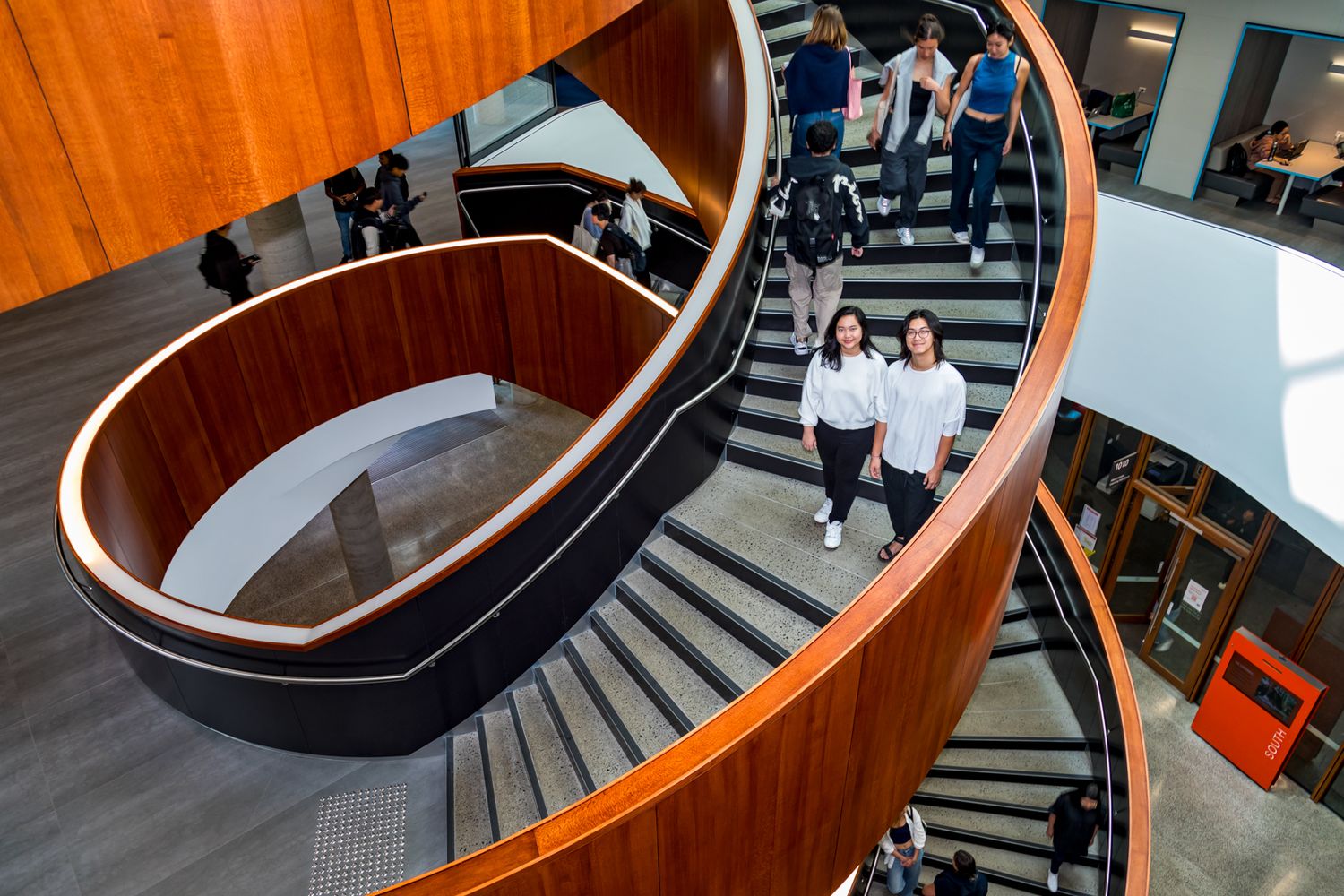 Overhead view of students walking up a sweeping wooden spiral staircase inside a modern multi-level university building.