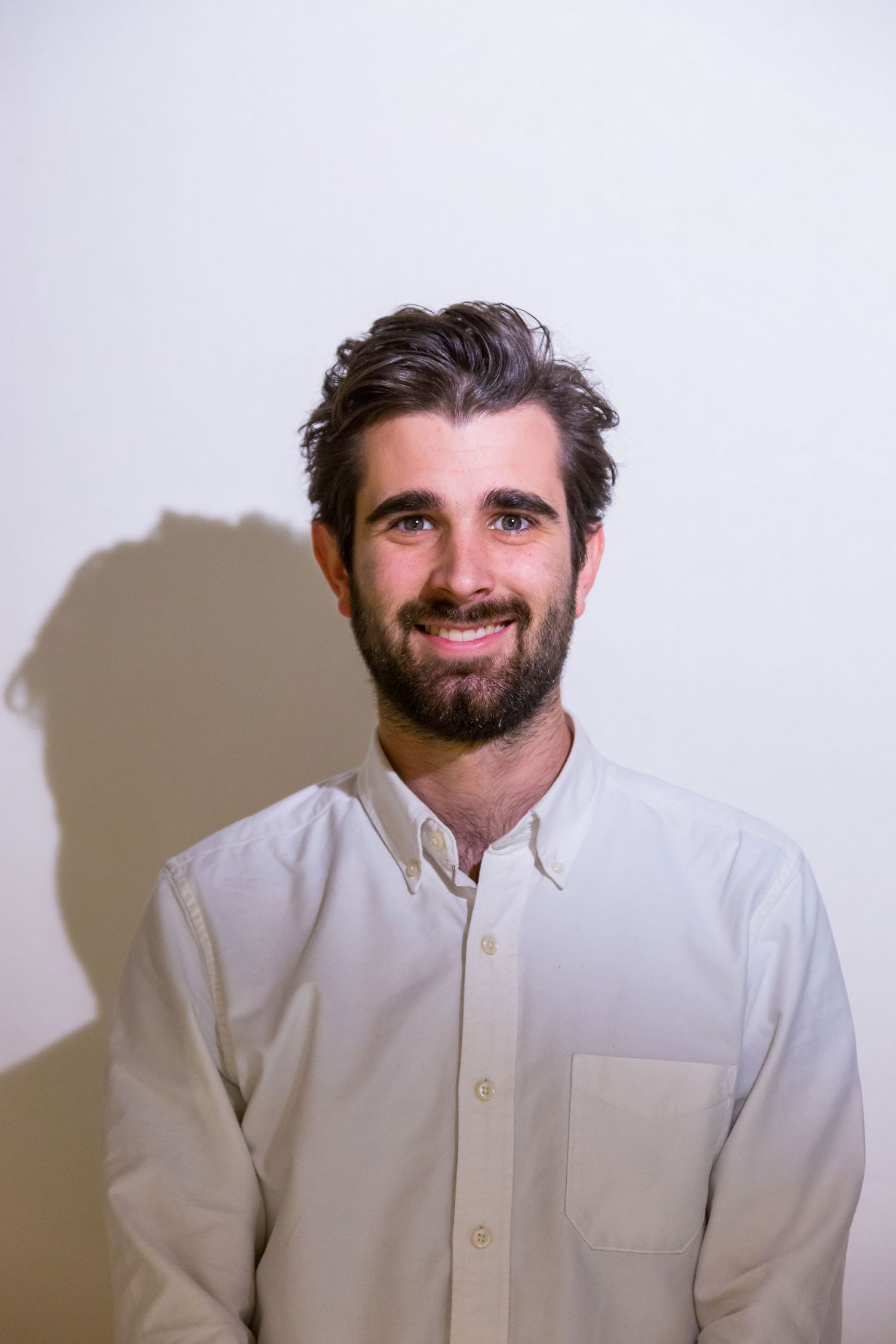 A smiling bearded man with dark tousled hair wears a white button-up shirt and poses against a plain light wall.