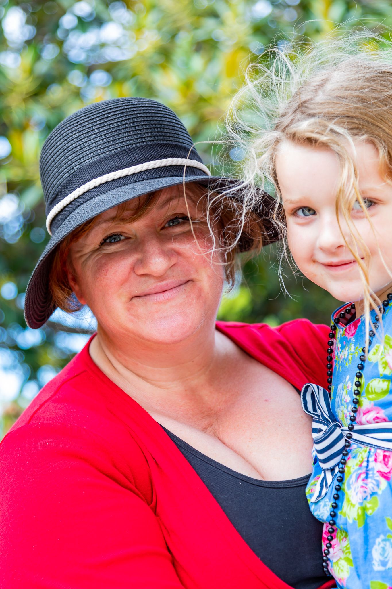 Smiling woman in a black sunhat and red cardigan holds a blonde young girl in a floral top outdoors among green foliage.