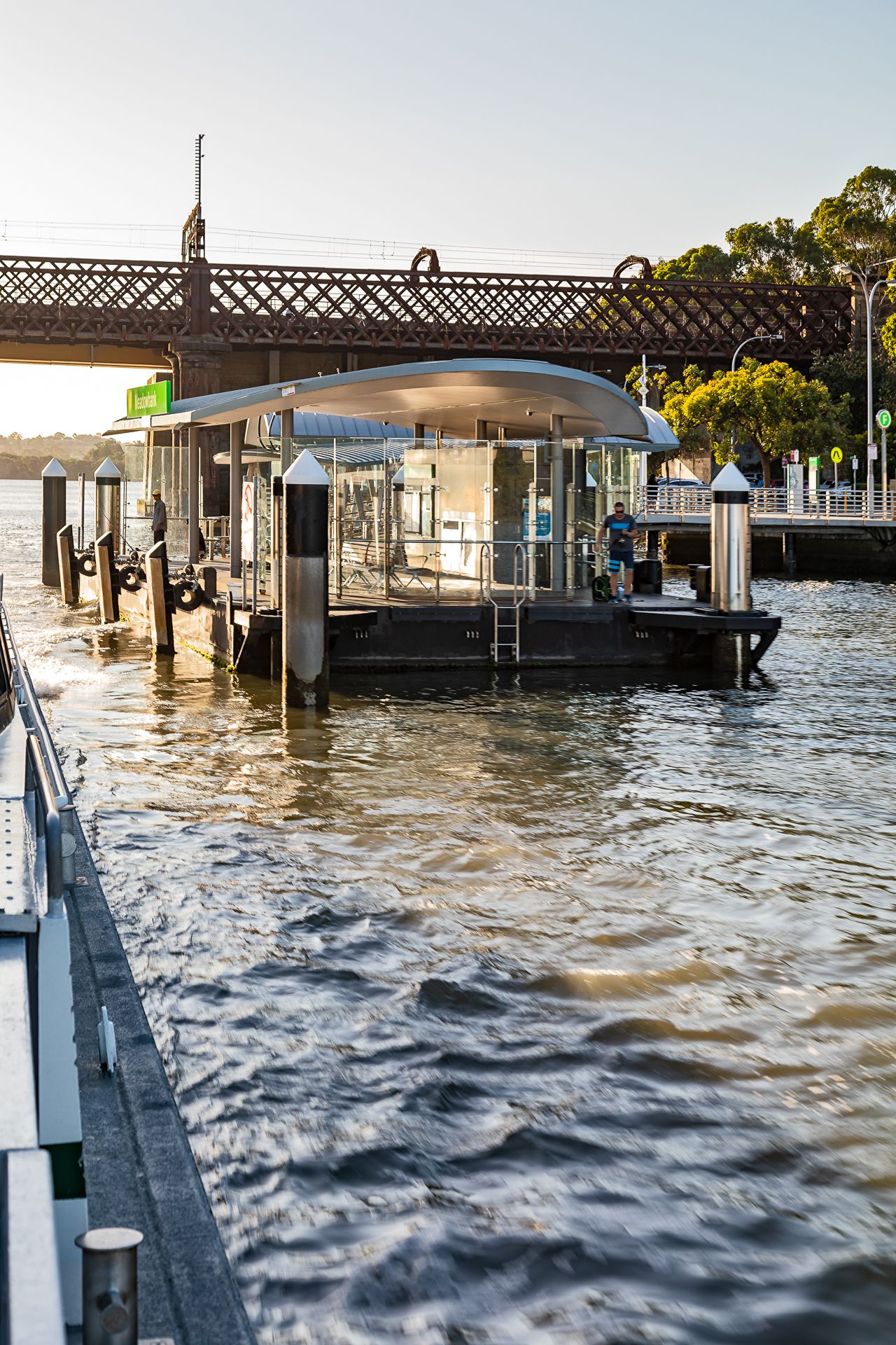 Ferry wharf with a curved roof on rippling river water, with a historic steel bridge and trees glowing in the evening light.