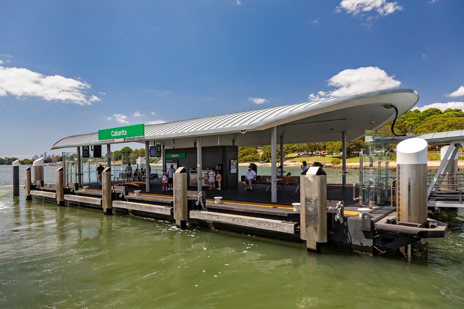 Ferry wharf with green sign and curved roof shelter beside moored pylons as passengers wait on the harbour waterfront.