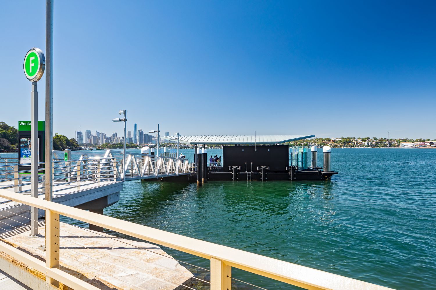 Ferry wharf pontoon with a parking sign and wooden railing overlooking blue harbour water and a distant city skyline.