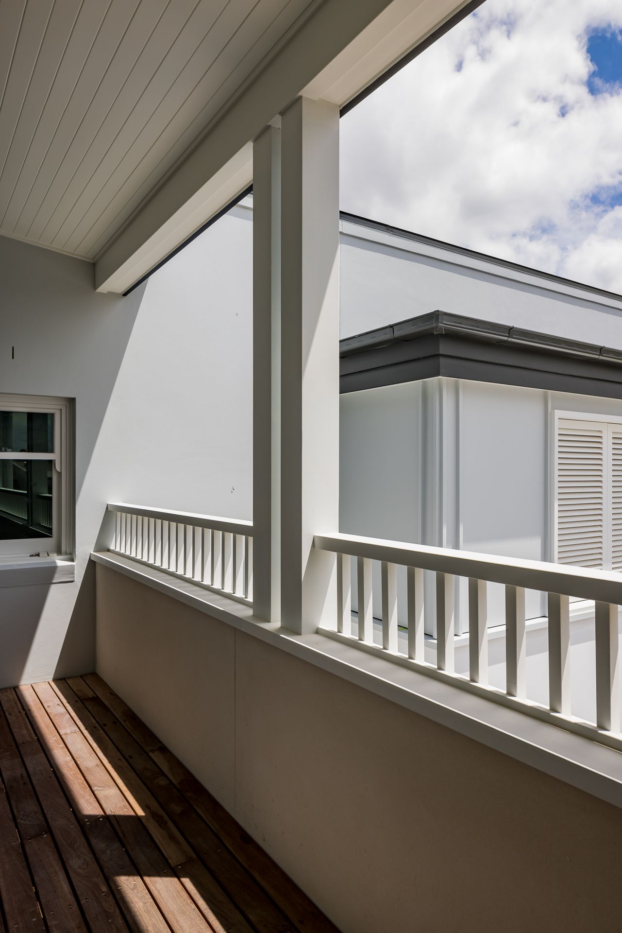 Sunlit timber-floored balcony with white balustrade and posts overlooking neighboring rooftops under a cloudy sky.