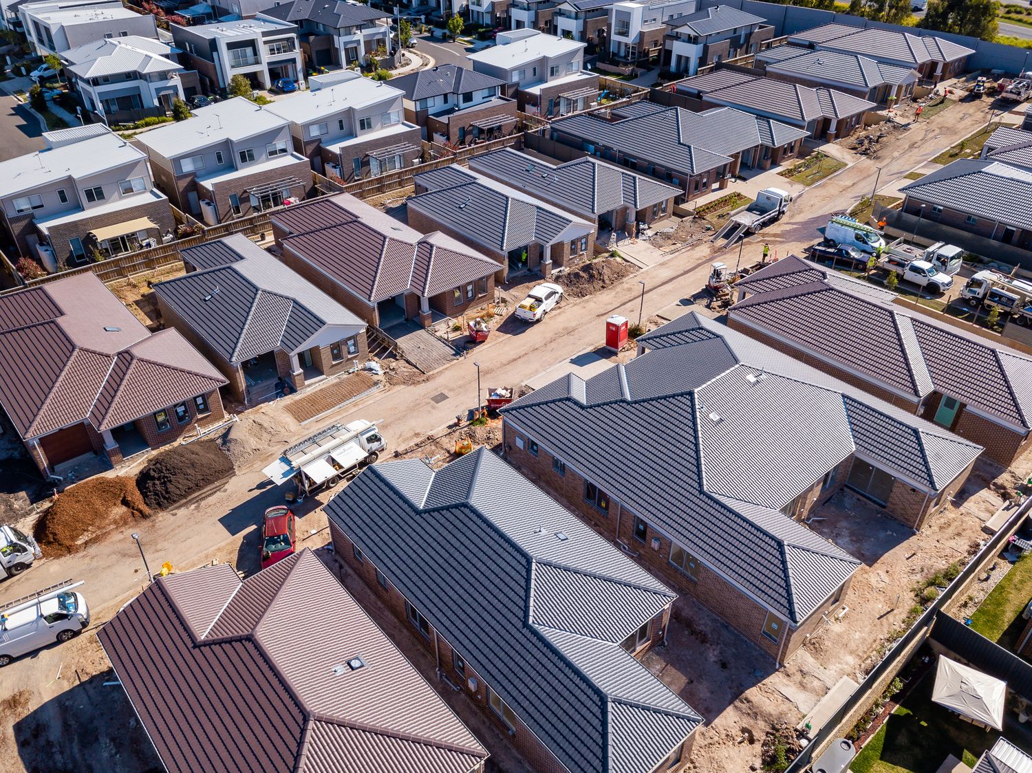 Aerial view of new houses under construction with tiled roofs, parked work vehicles and dirt lots in a growing suburban estate.