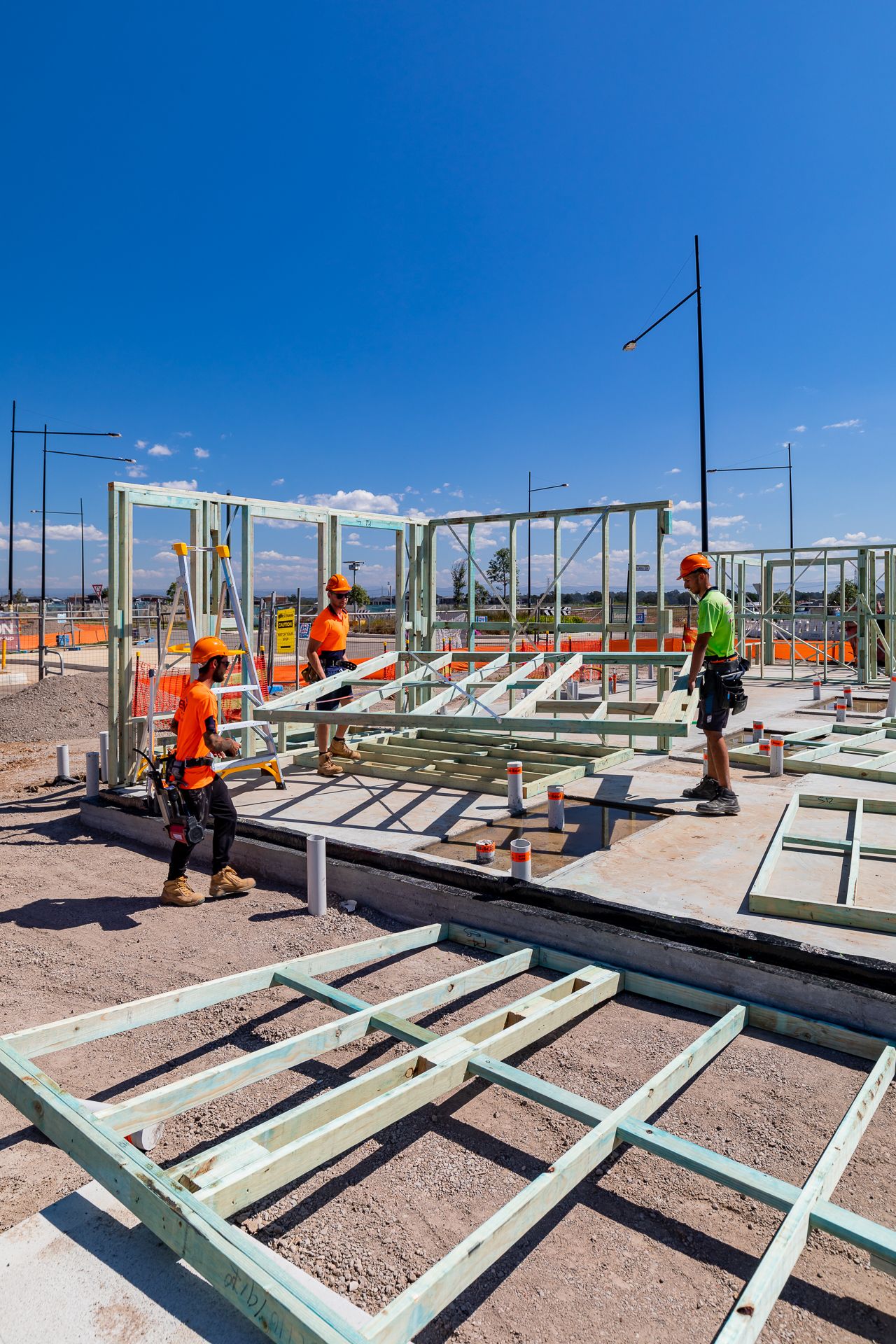 Three builders in orange hard hats and safety gear erect green steel wall frames on a slab under a clear blue sky.