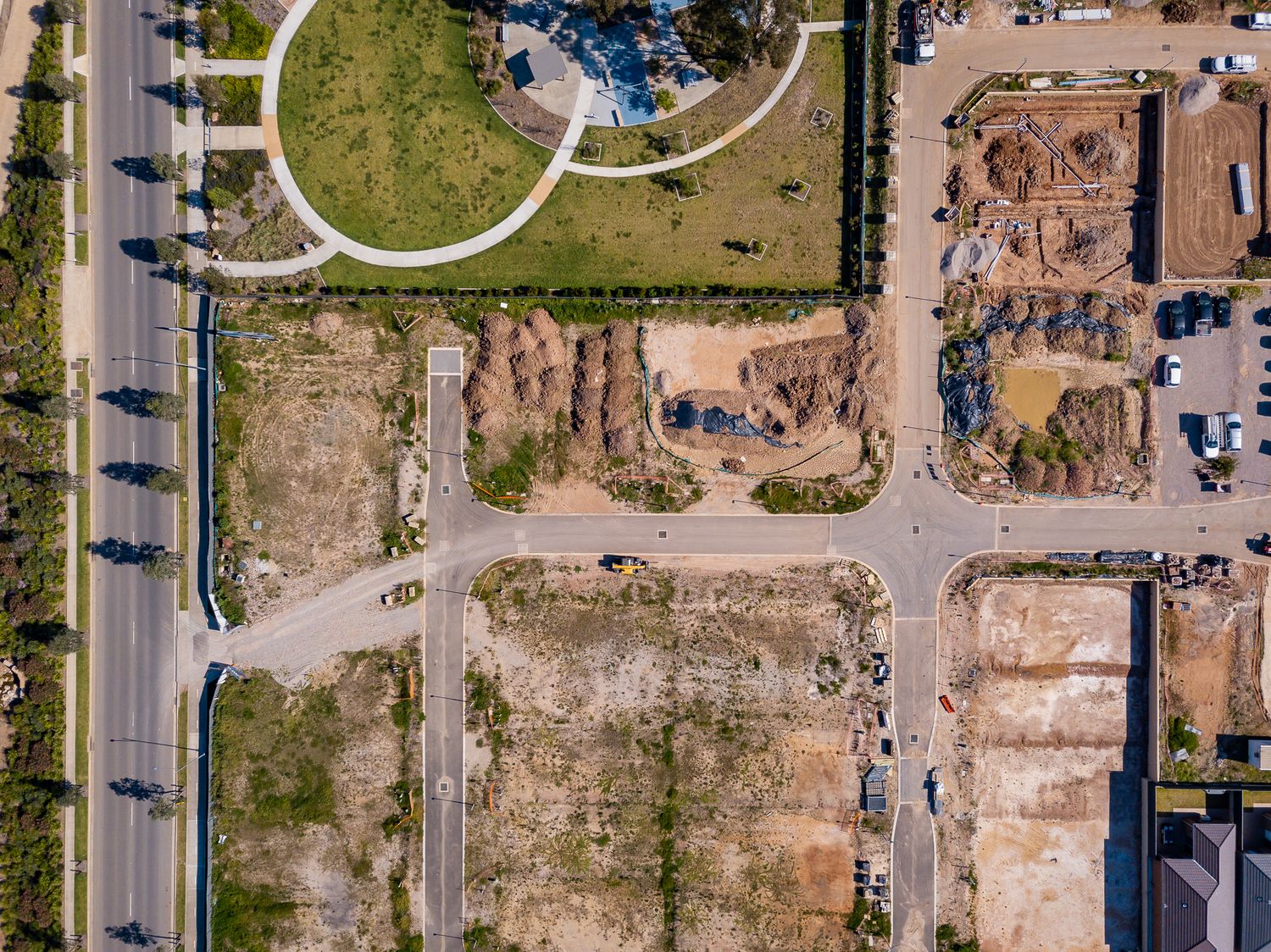 Overhead drone shot of a new suburban estate with vacant building lots, dirt roads, a curved park path and a tree-lined main road.