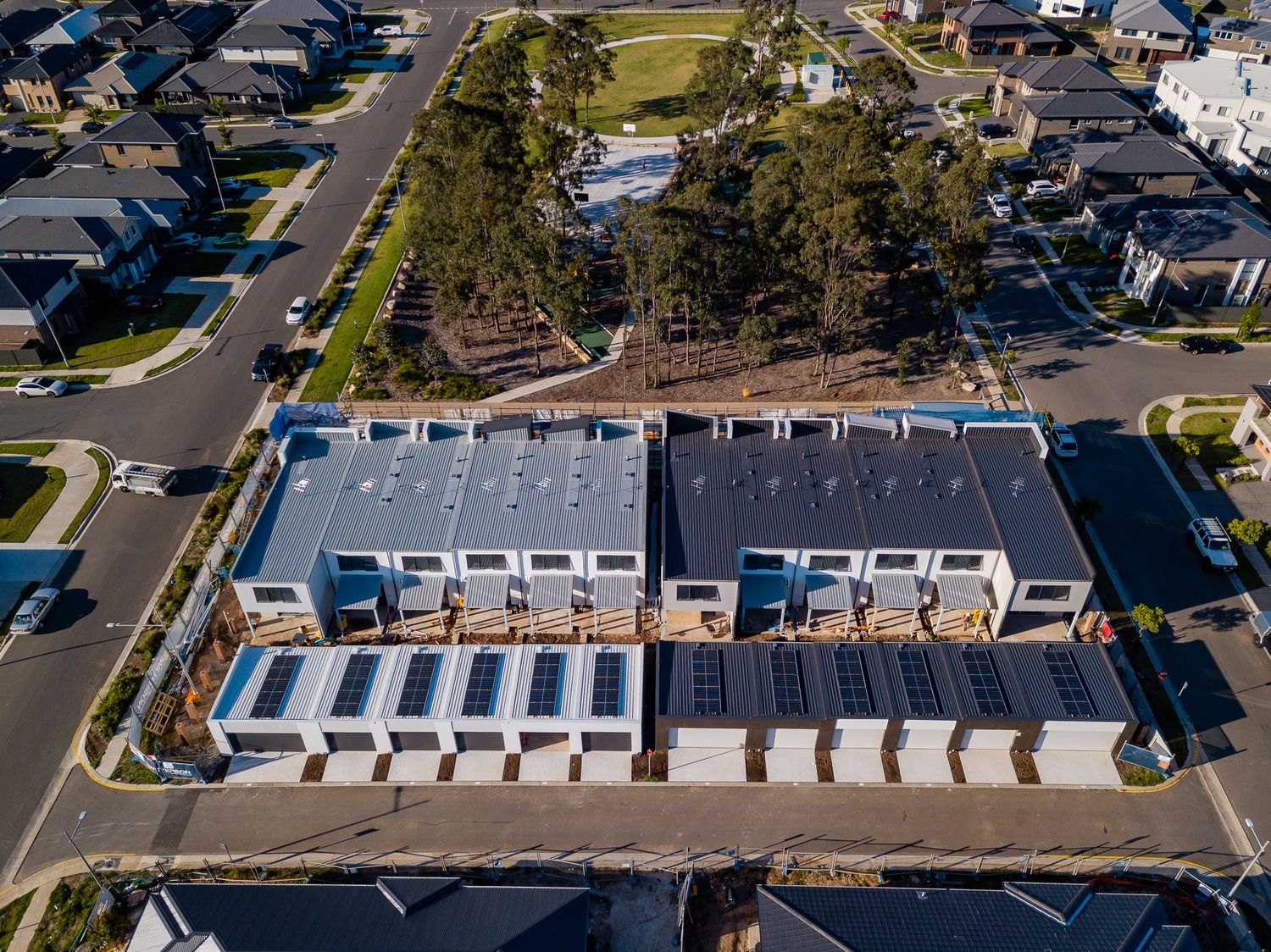 Aerial view of modern townhouses with grey roofs and rooftop solar panels in a new suburban estate with tree-lined streets and parkland.