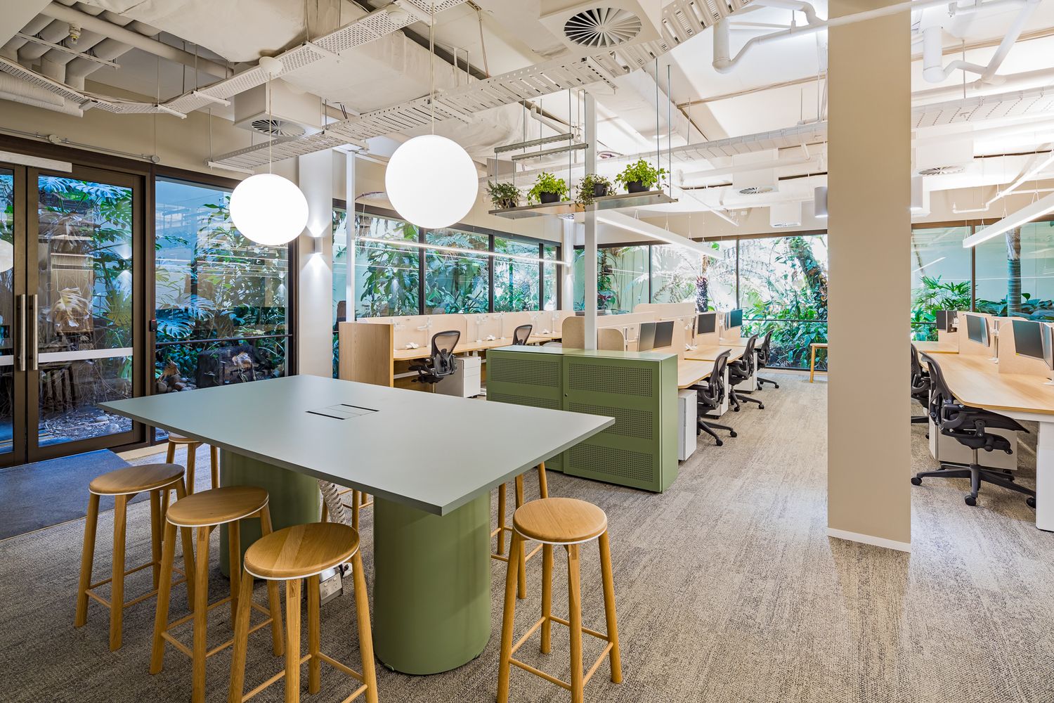 Modern open-plan office with a large communal table, wooden stools, green desk partitions and floor-to-ceiling windows overlooking greenery.