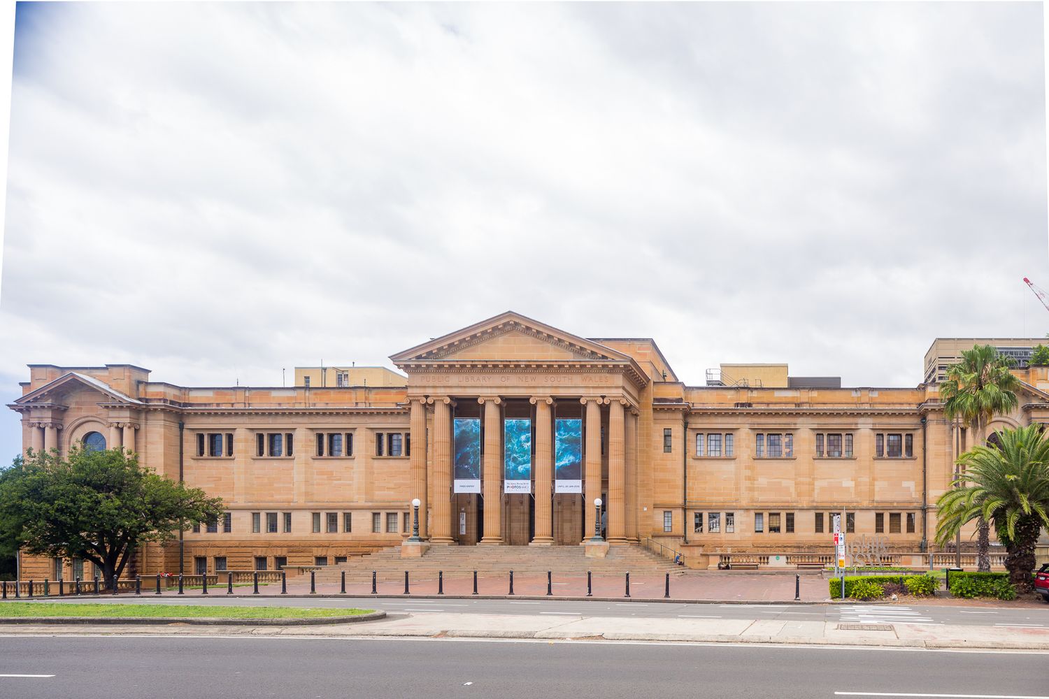 Grand neoclassical sandstone library building with a columned portico and blue banners under a cloudy sky, with palm trees at the sides.