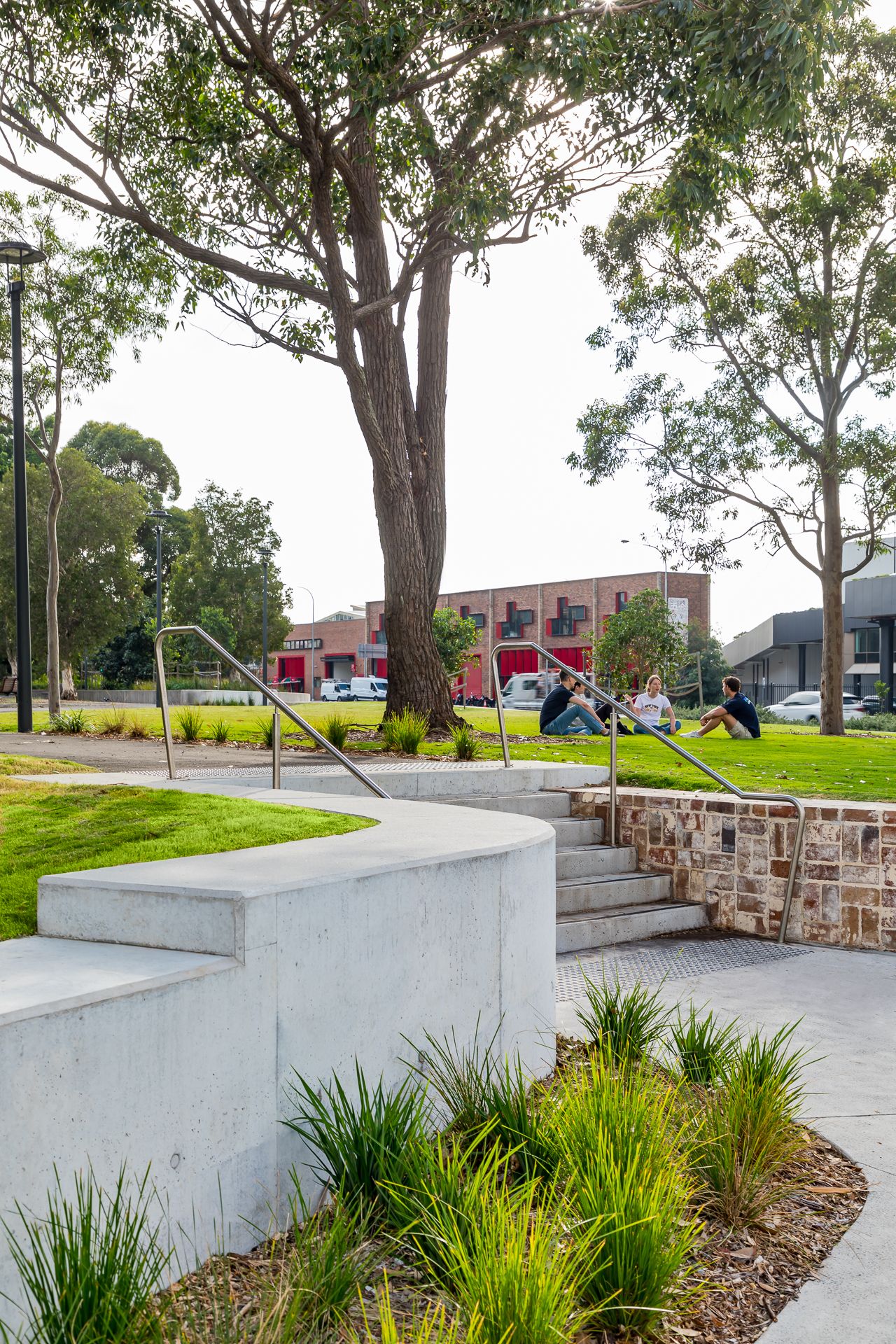 Curved concrete retaining wall and stone steps in a park, with people relaxing on grass near a tall tree and brick building.