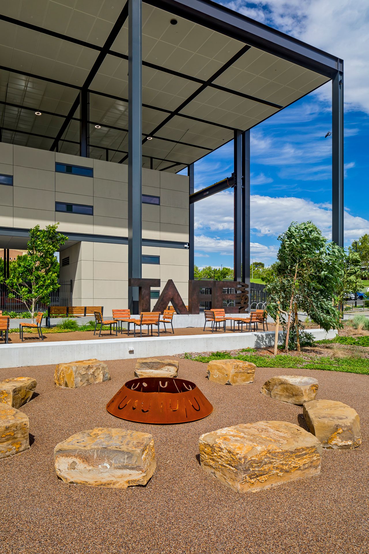 Outdoor campus courtyard with sandstone boulder seating around a rust-colored fire pit, timber benches and a covered steel walkway.