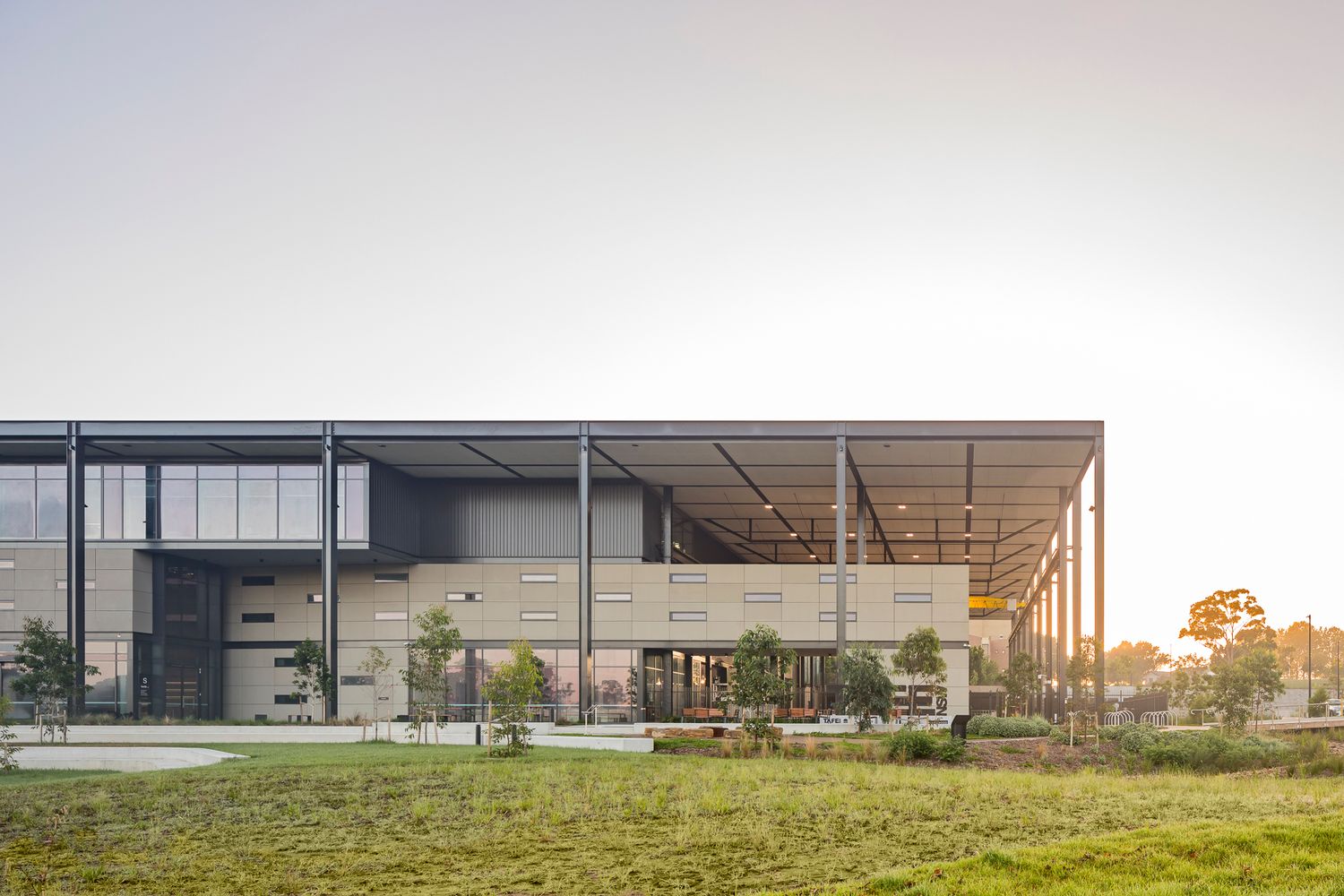 Front view of a modern steel-framed building with glass facade, stone panels and a large roof overhang above a green lawn.