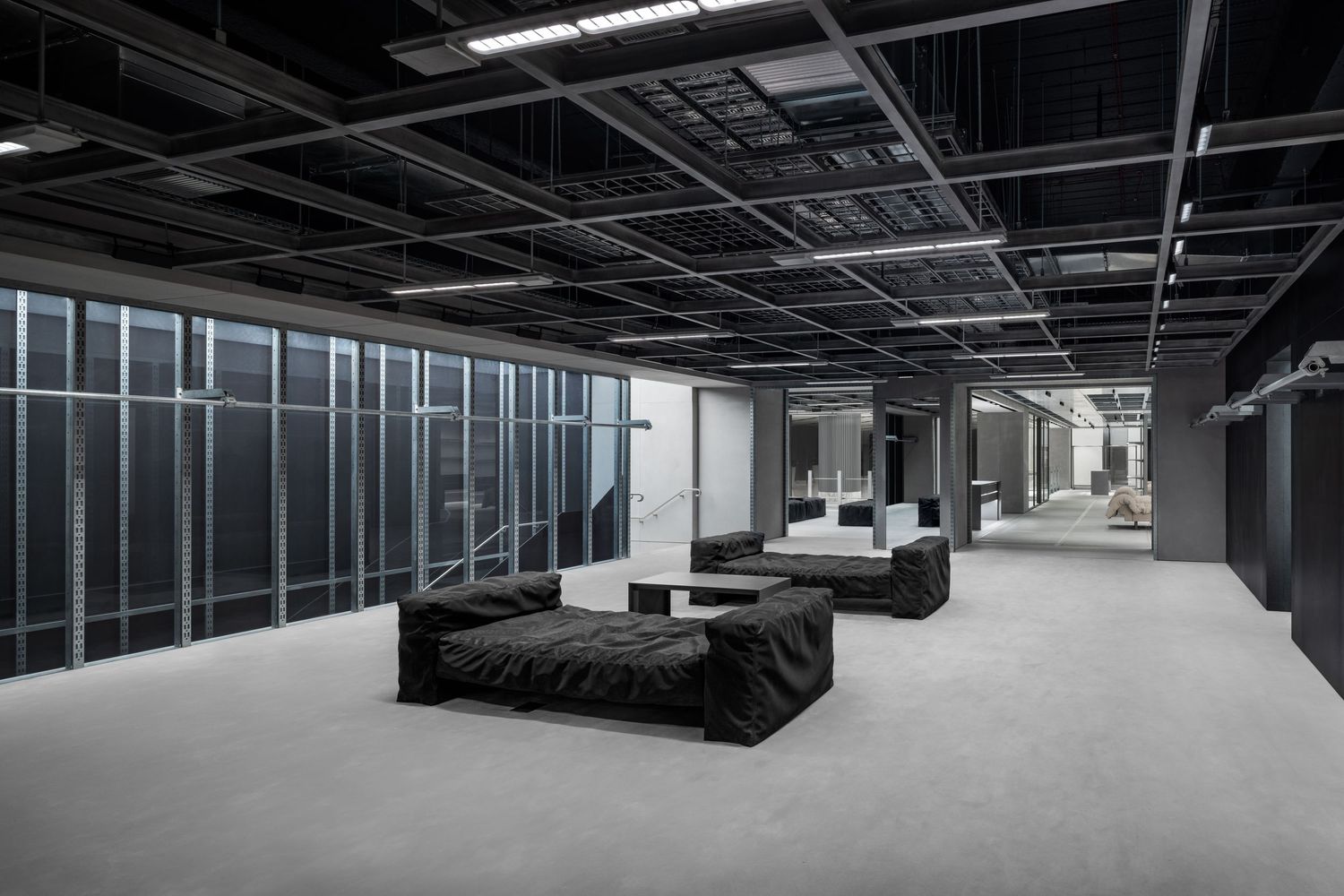 Minimalist industrial interior with black ceiling grid, floor-to-ceiling glass walls, and dark modular sofas on a gray concrete floor.