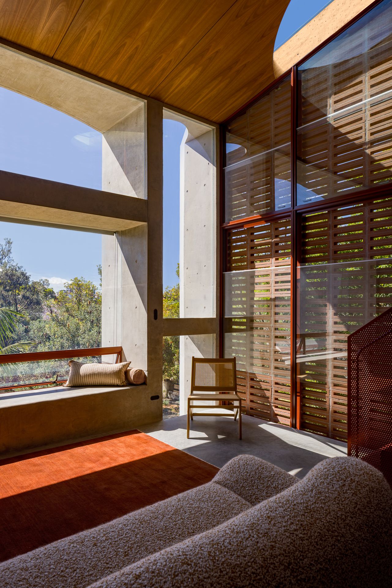 Bright living area with a concrete window seat, timber ceiling, louvred glazing, rust-red rug, wooden chair and bouclé sofa.