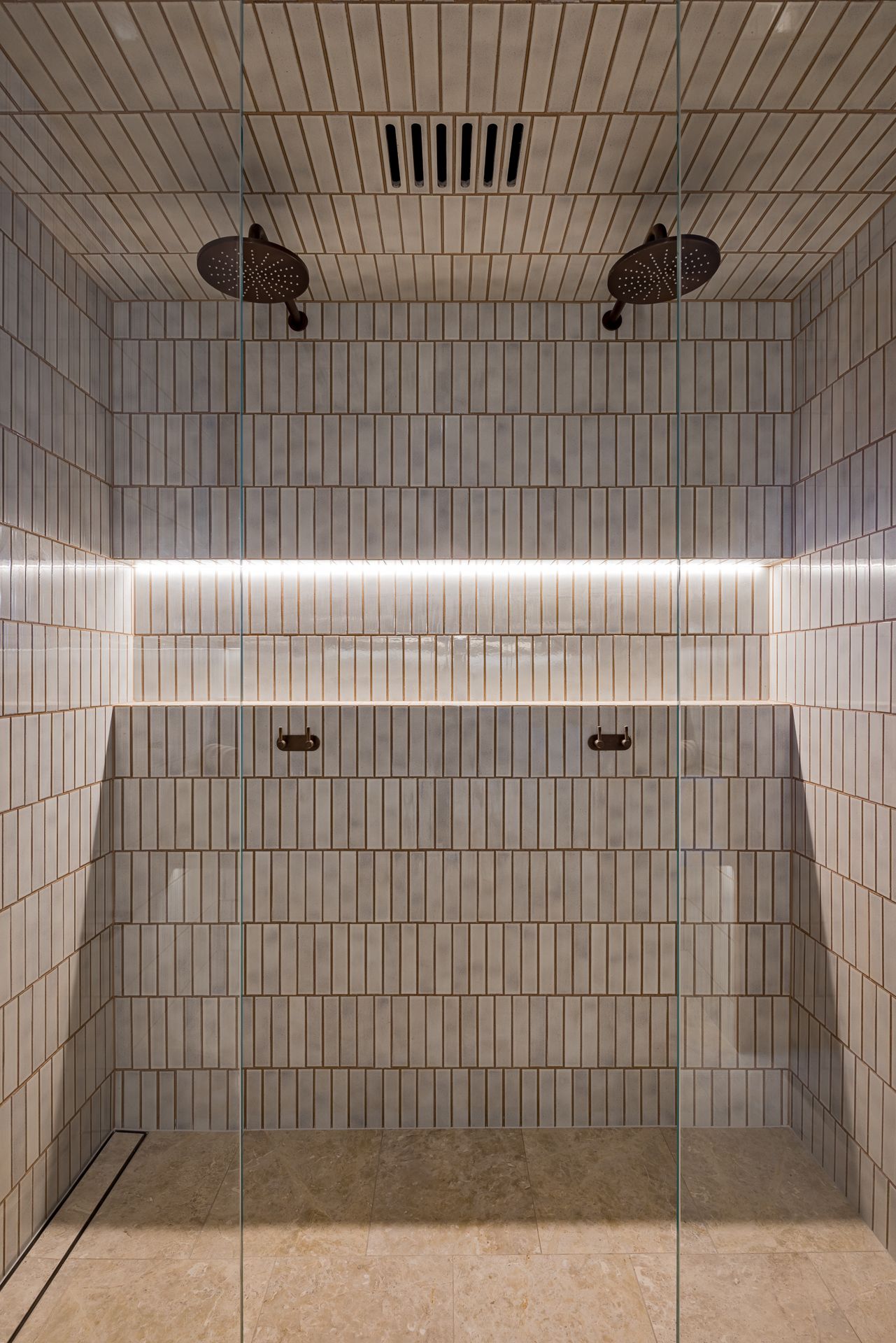 Walk-in shower with vertical white tiles, twin round rainfall showerheads, a recessed LED-lit niche, and a glass enclosure.