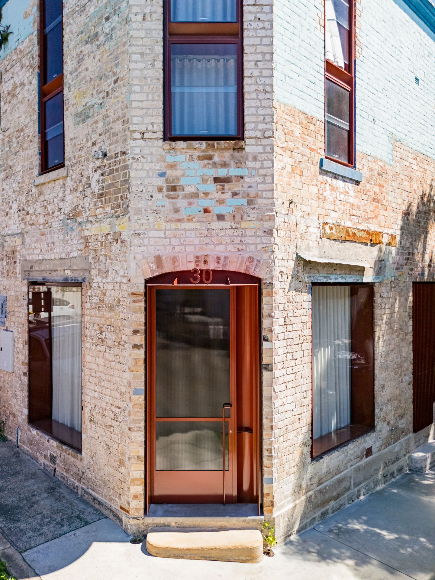 Corner of a heritage brick building with a rust-red timber door numbered 30, arched brick lintel, and tall timber-framed windows.