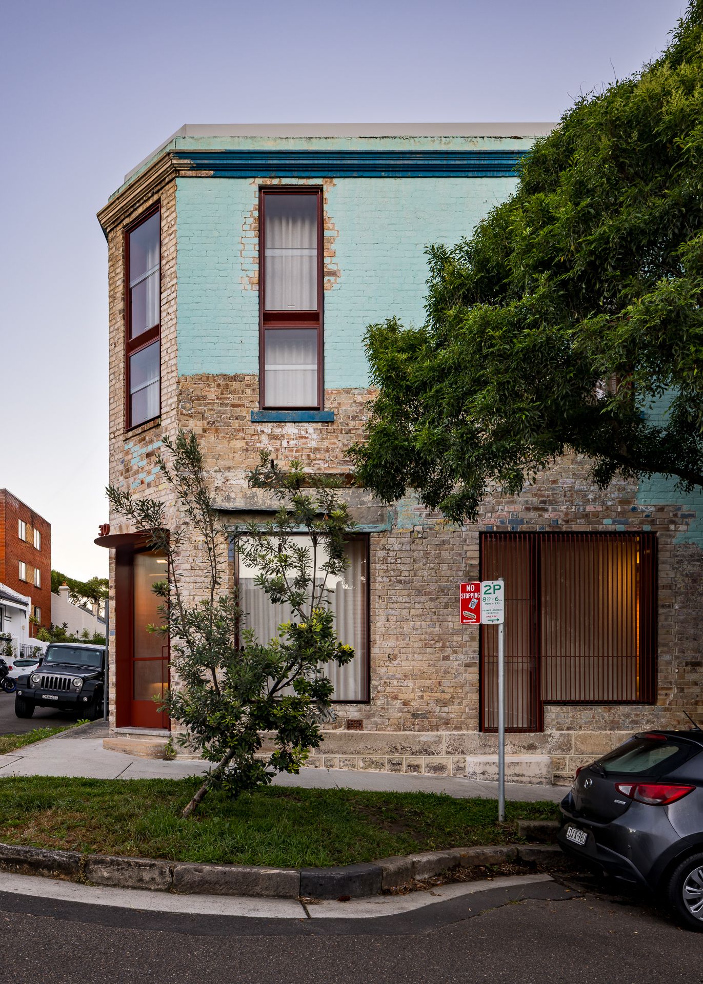 Renovated heritage brick terrace on a street corner with teal upper walls, burgundy window frames and a parked car nearby.