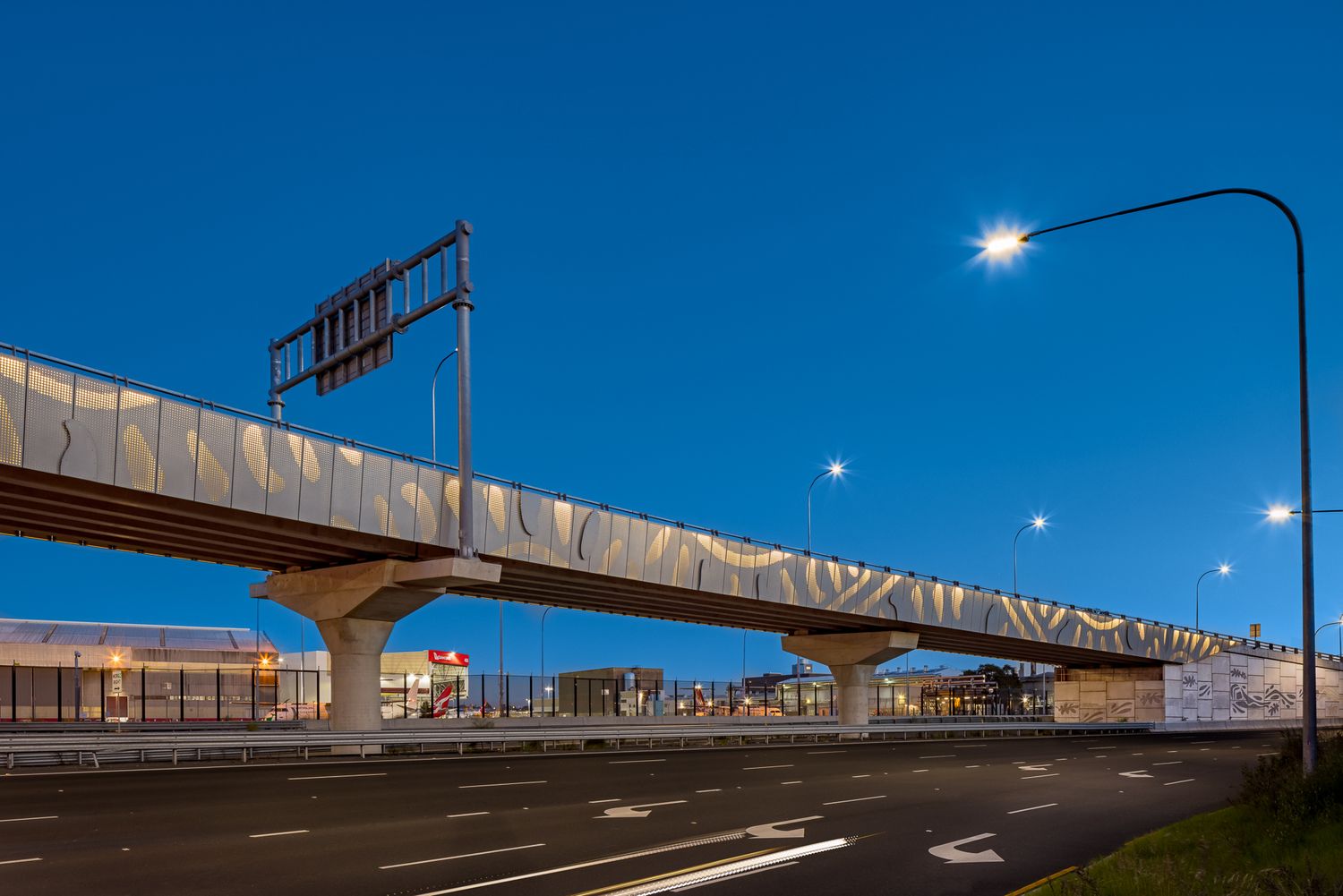 Elevated viaduct with textured silver cladding and angular concrete supports crosses a multi-lane road under a clear twilight sky.