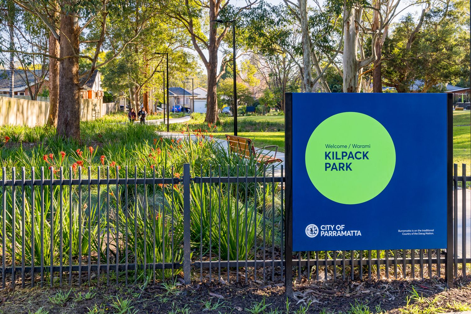 Blue welcome sign for Kilpack Park, City of Parramatta, beside a black fence and lush native garden with a path and trees.