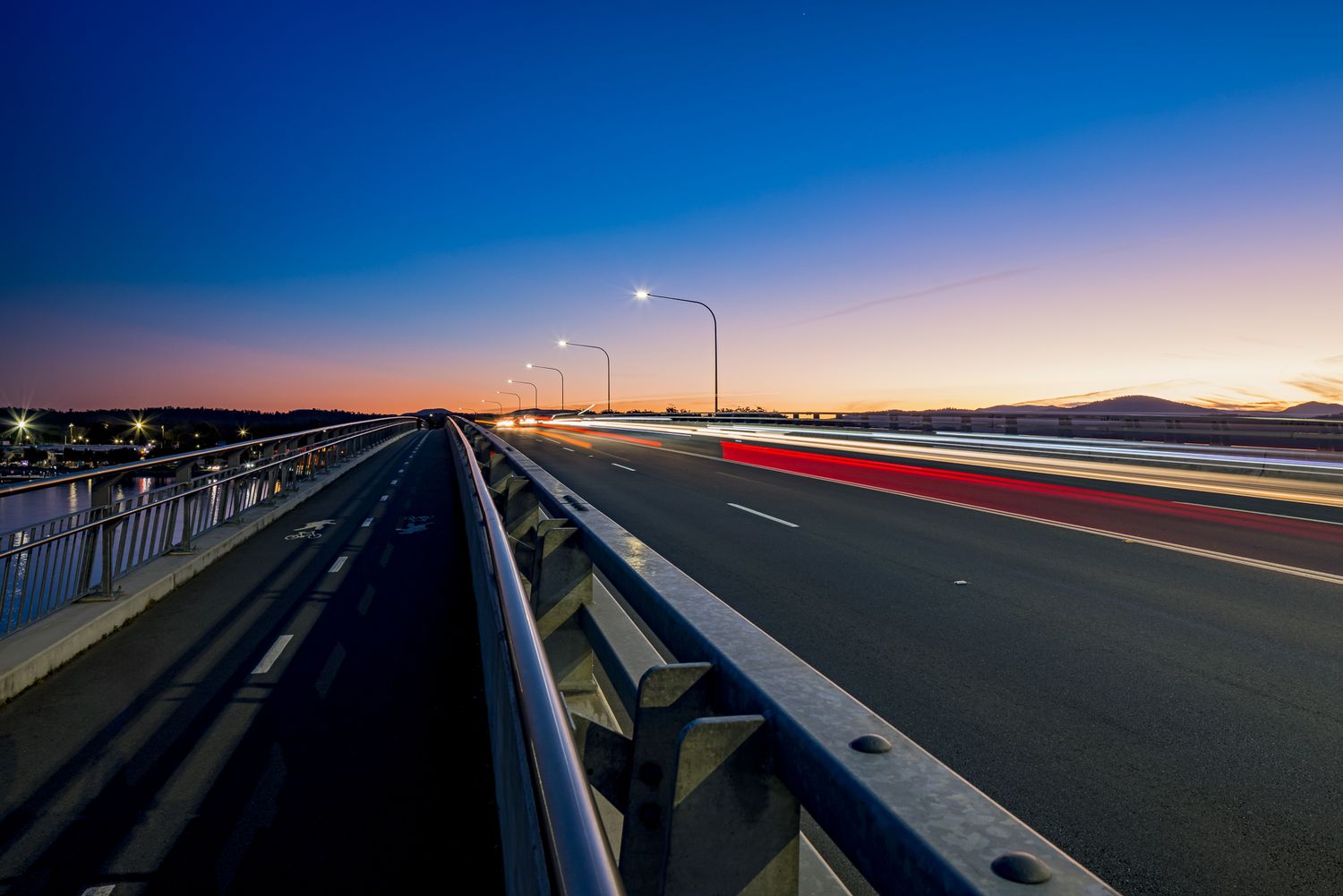 Bridge roadway with light trails and street lamps at dusk, with a glowing orange and purple sky over the horizon.